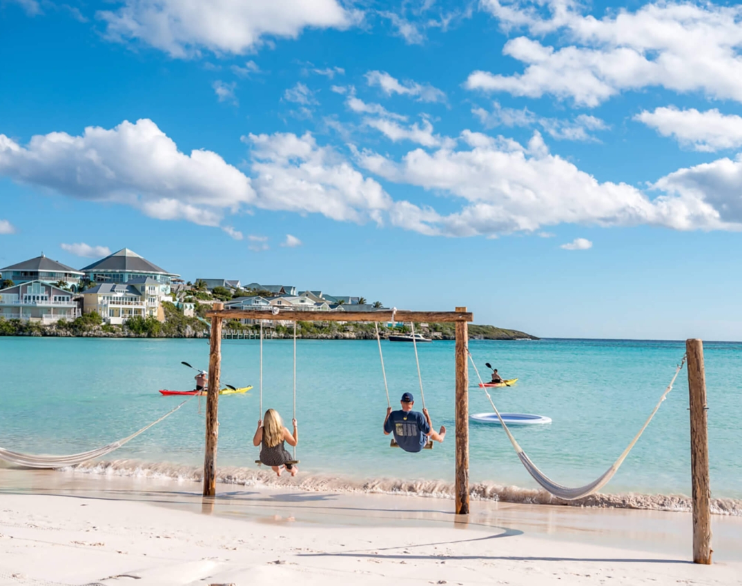 Two people on wooden swings at The Abaco Club offering a leisurely club lifestyle amidst the stunning coastal landscape of the Bahamas.