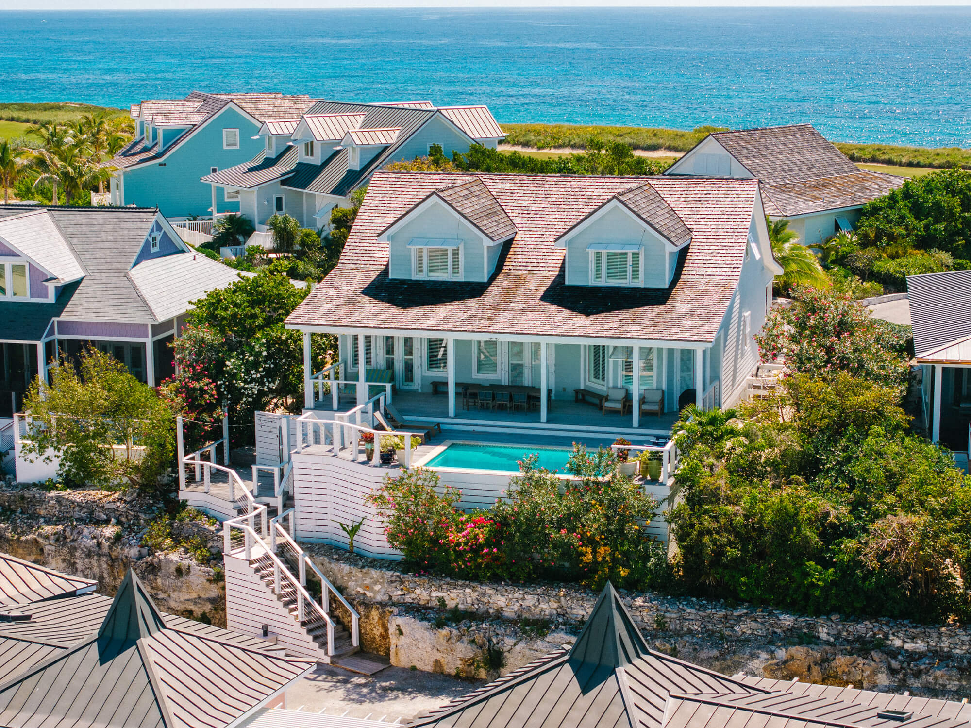 Aerial view of blue seaside cottages with white trim and brown roofs along the coast, pool in foreground and turquoise ocean beyond.