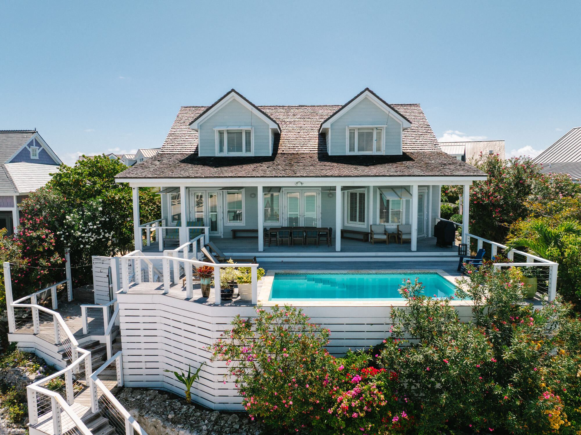 Two-dormer blue house with white trim, deck and a rectangular pool in front among flowering shrubs