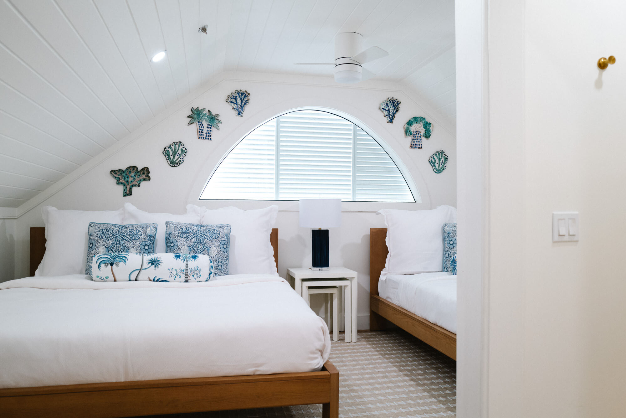 Twin white-bedroom with wooden bed frames, blue patterned cushions, and a curved window framed by coastal ceramic wall art on each side.