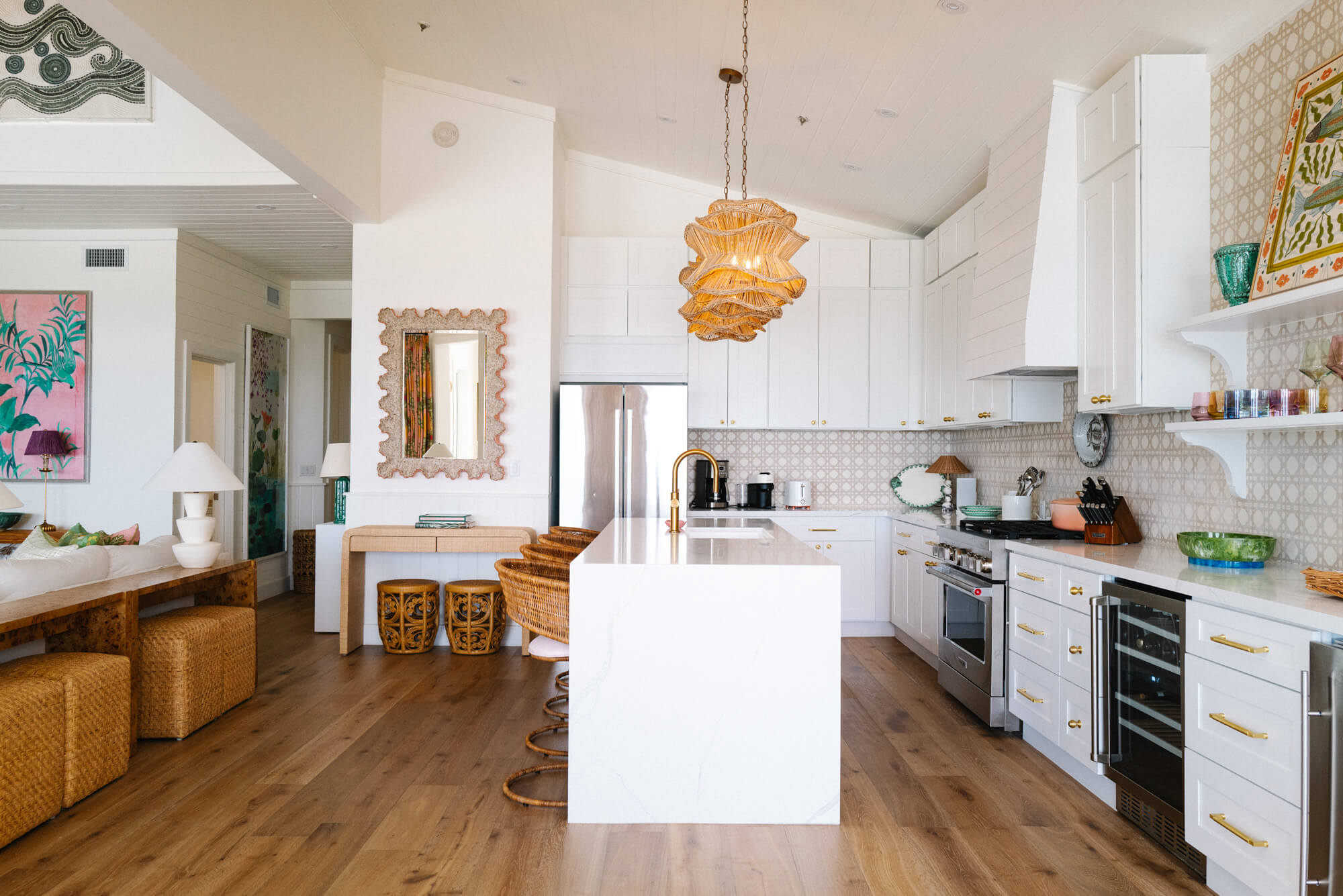 Bright open-plan kitchen with white cabinets, a marble island, and woven pendant light centerpiece; wicker stools line the island and wooden floors run throughout.