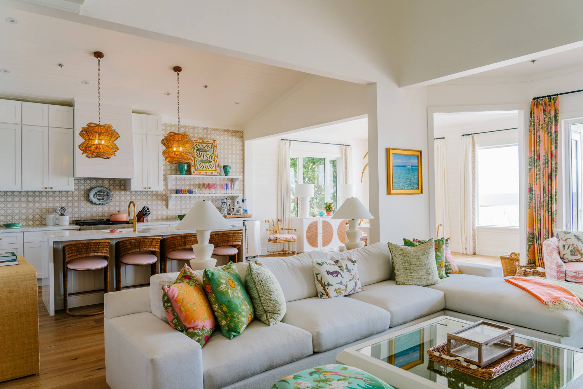 Open-concept kitchen and living room with white cabinets, two woven pendant lights, and a gray sectional with tropical-patterned cushions.