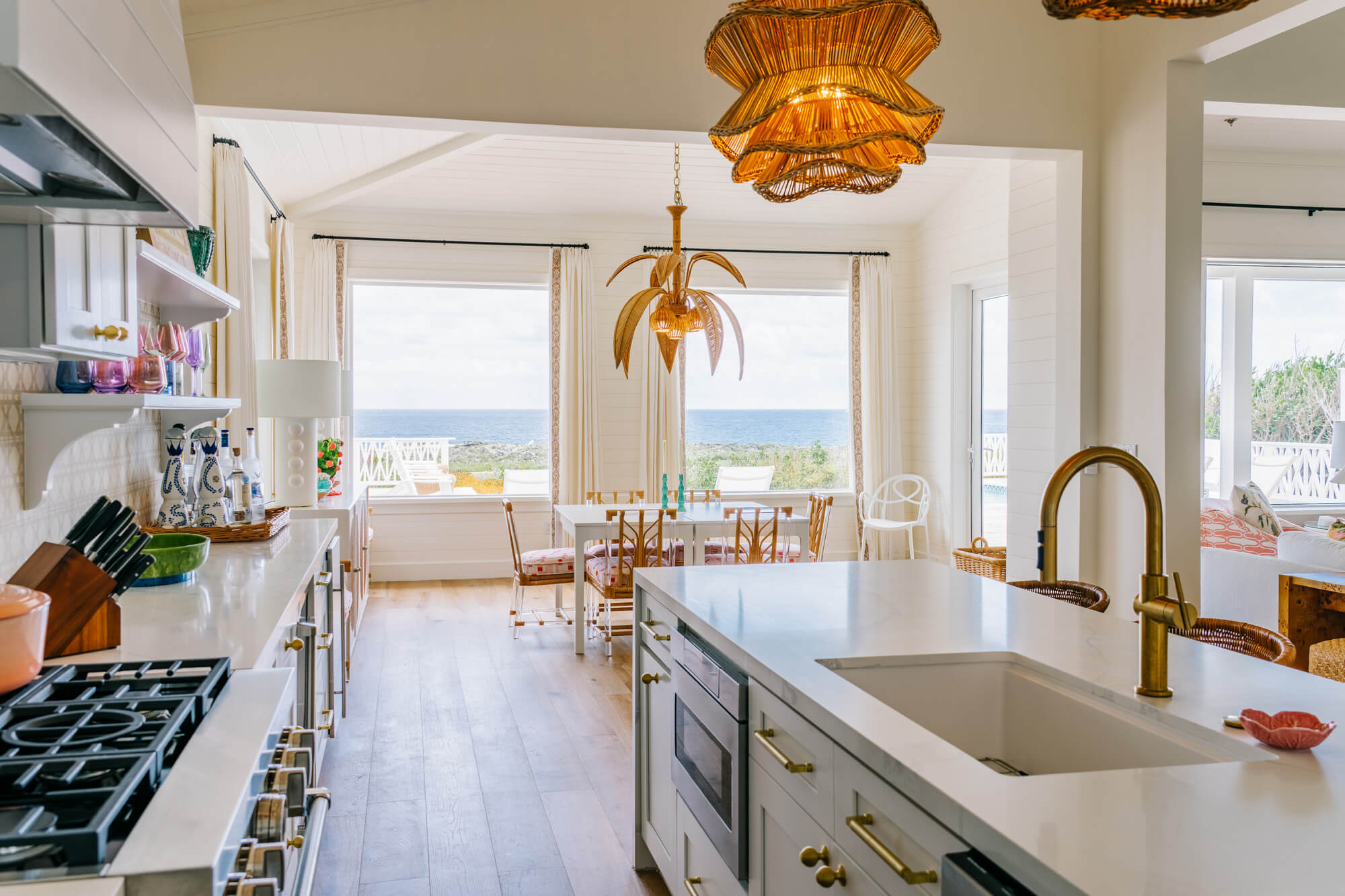 Bright coastal kitchen with white cabinets, brass hardware, large island, pendant wicker lights, and ocean view through windows.