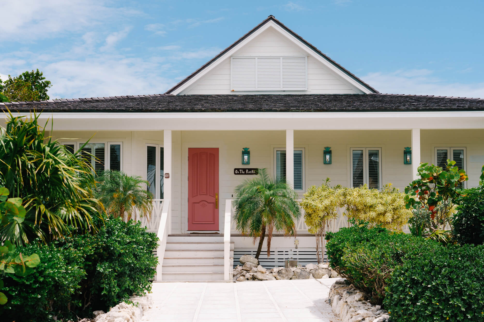 Front view of a white bungalow with a red front door, covered porch, and lush tropical plants lining a stone path.