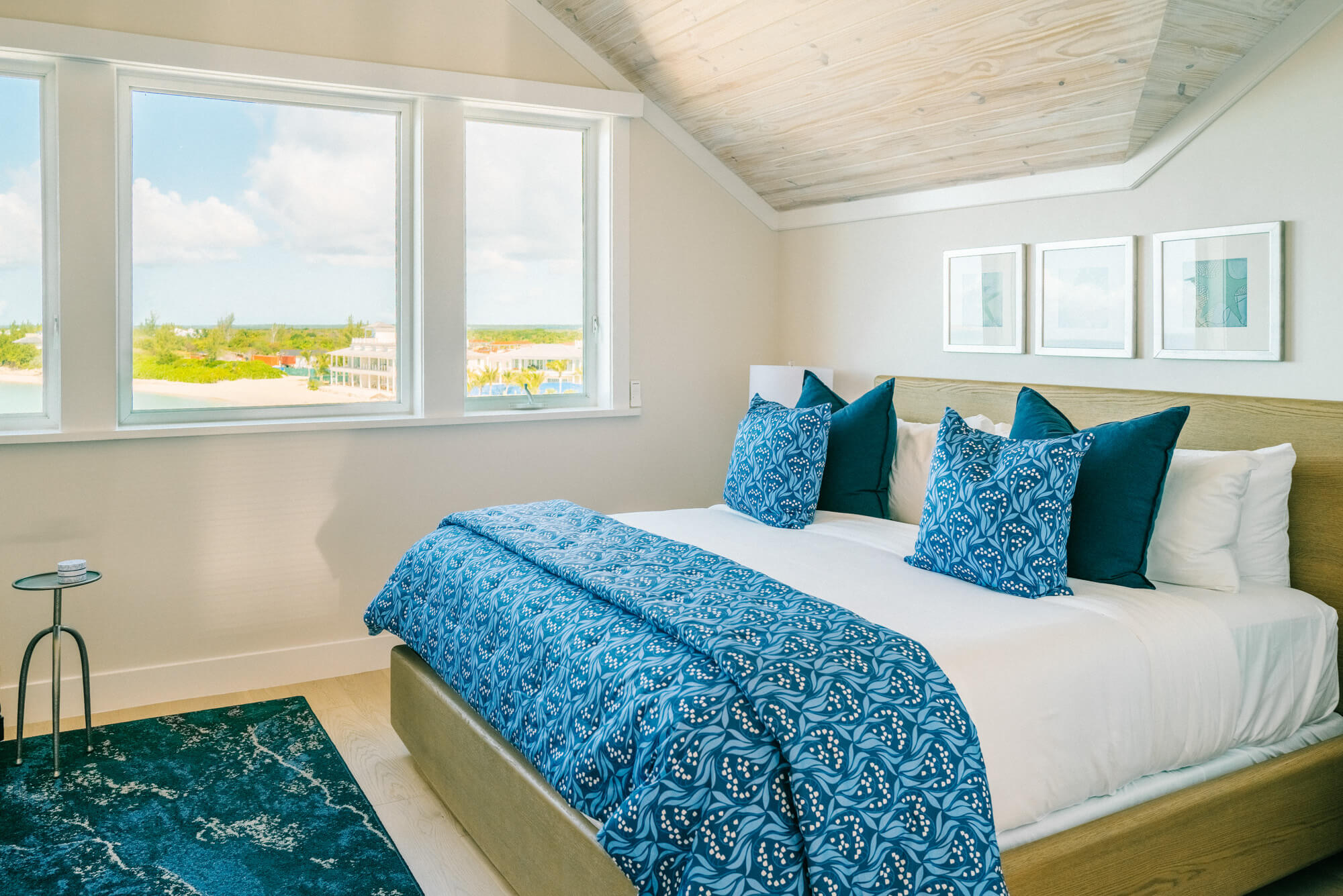 Bedroom with a sea view: white walls, large windows, and blue patterned bedding on a low bed with teal pillows.