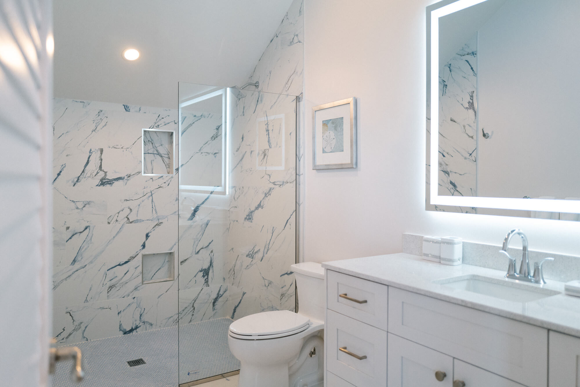 Modern bathroom with marble-tiled shower, glass door, white vanity, and backlit rectangular mirror