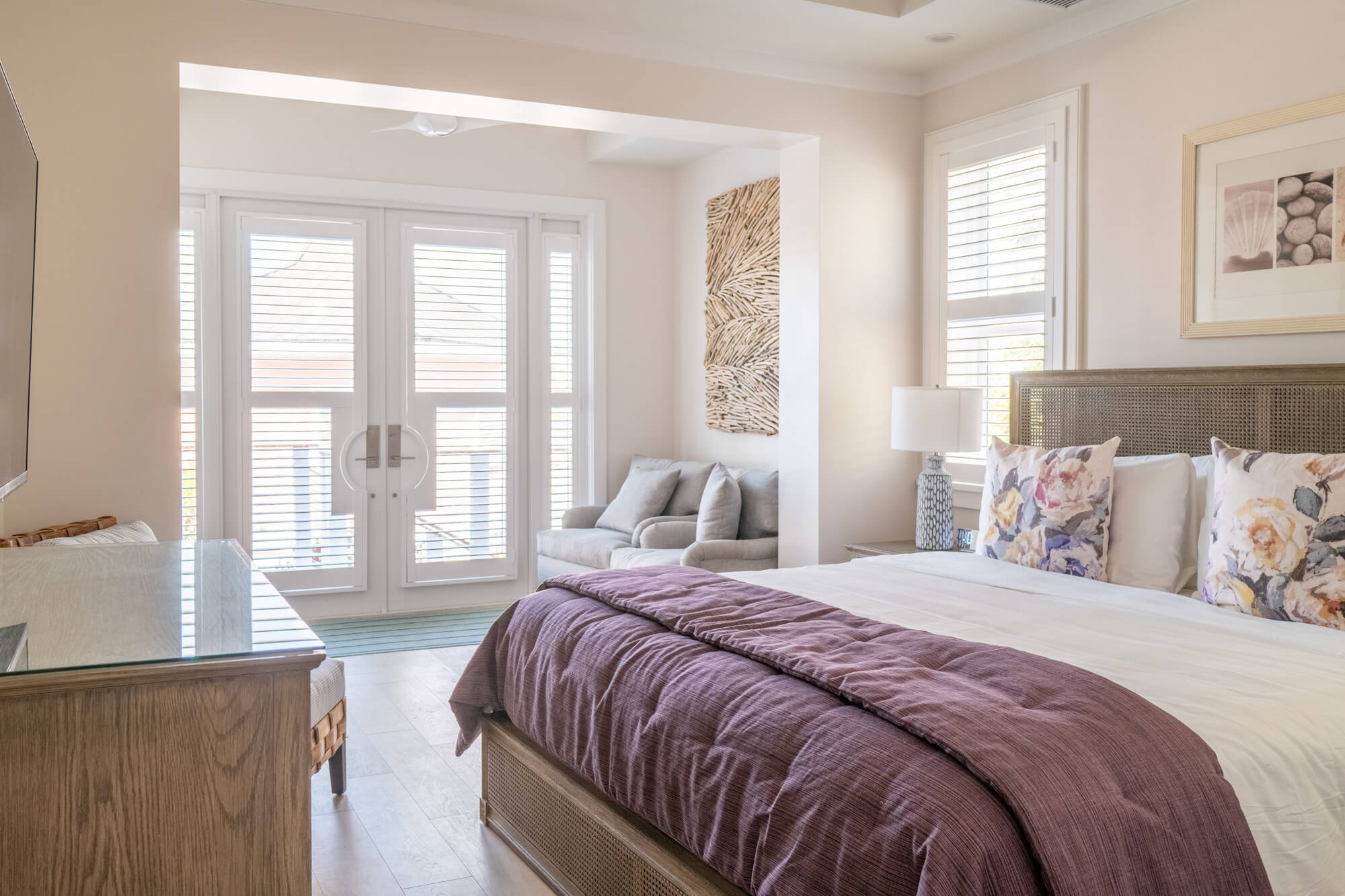 Bright hotel bedroom with two beds, purple duvets, floral throw pillows, and a wicker headboard near glass French doors with blinds.