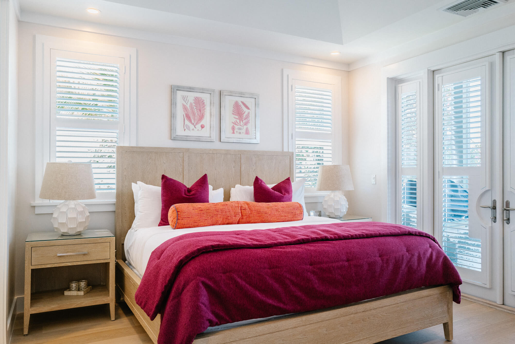 Bedroom with a wooden platform bed, magenta duvet and accent pillows, two white lamps on glass-topped nightstands, white shutters behind.