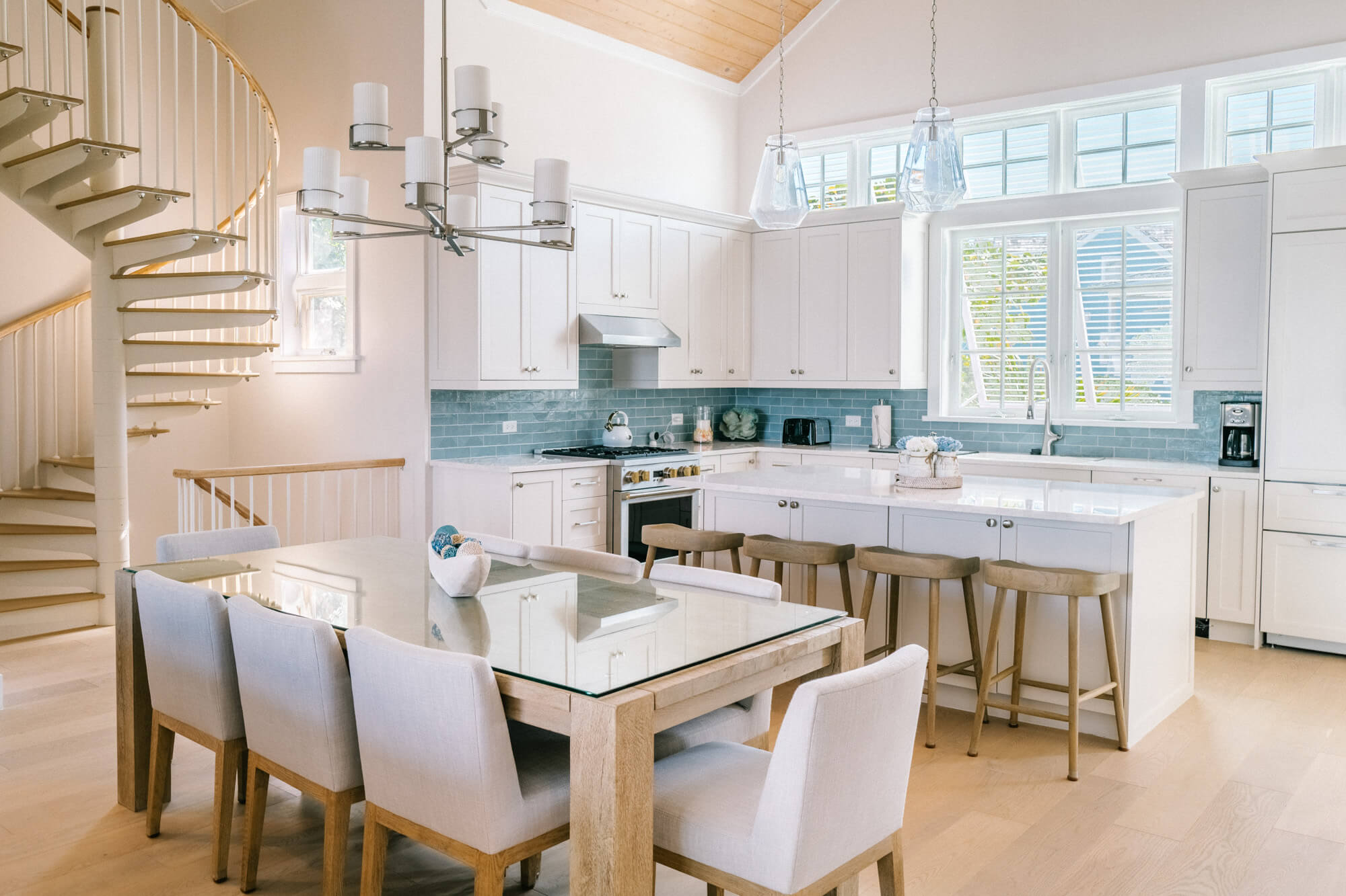 Bright open-concept kitchen with white cabinets, turquoise backsplash, and large windows; wooden spiral staircase on the left.