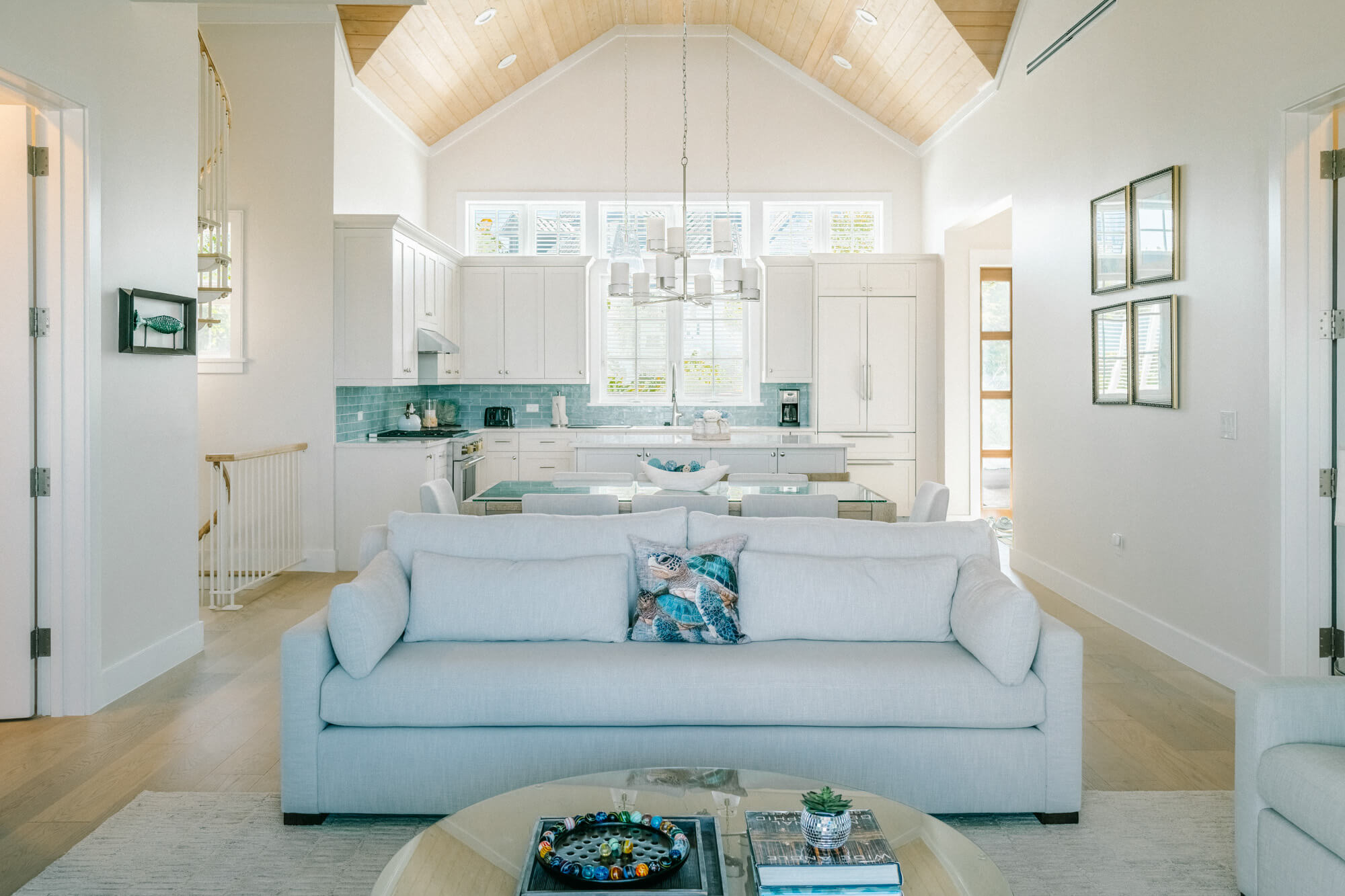 Open-concept living area with a light blue sofa facing a glass coffee table; a bright kitchen with white cabinets, turquoise tile backsplash, and a vaulted wooden ceiling in the background.
