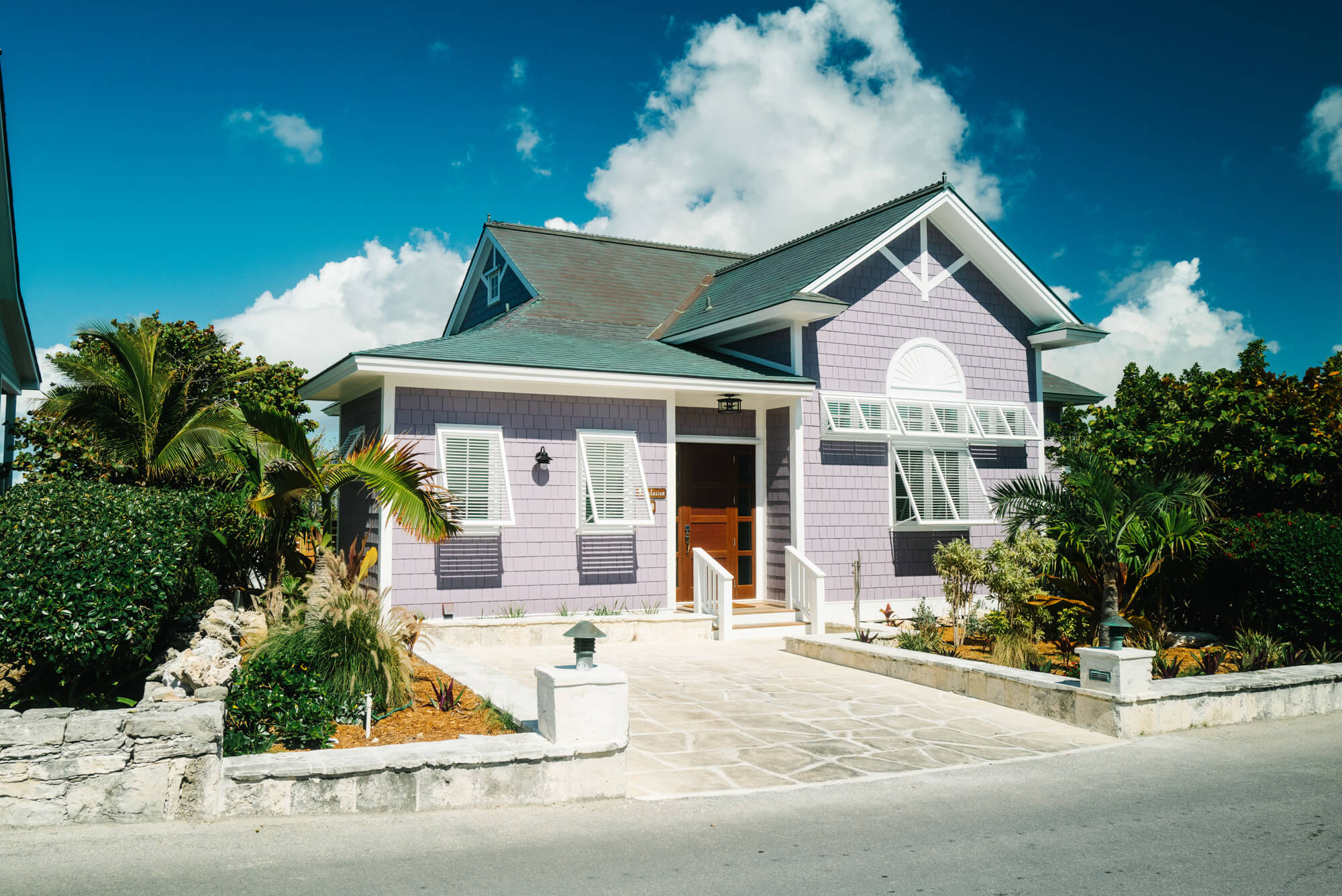 Lavender-colored house with white trim and gabled roof, front steps leading to wooden door, tropical plants framing the entry under a bright blue sky.