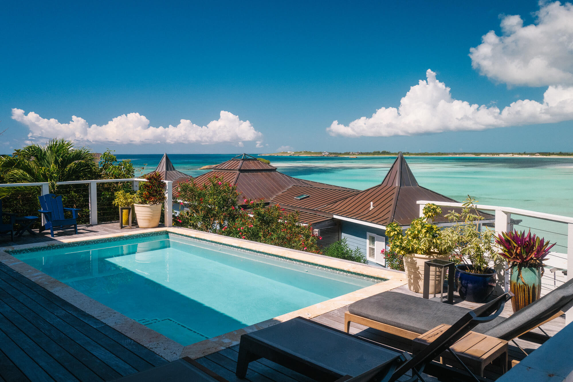 Poolside terrace with blue lounge chairs and potted plants, overlooking a turquoise ocean under a blue sky with fluffy clouds.