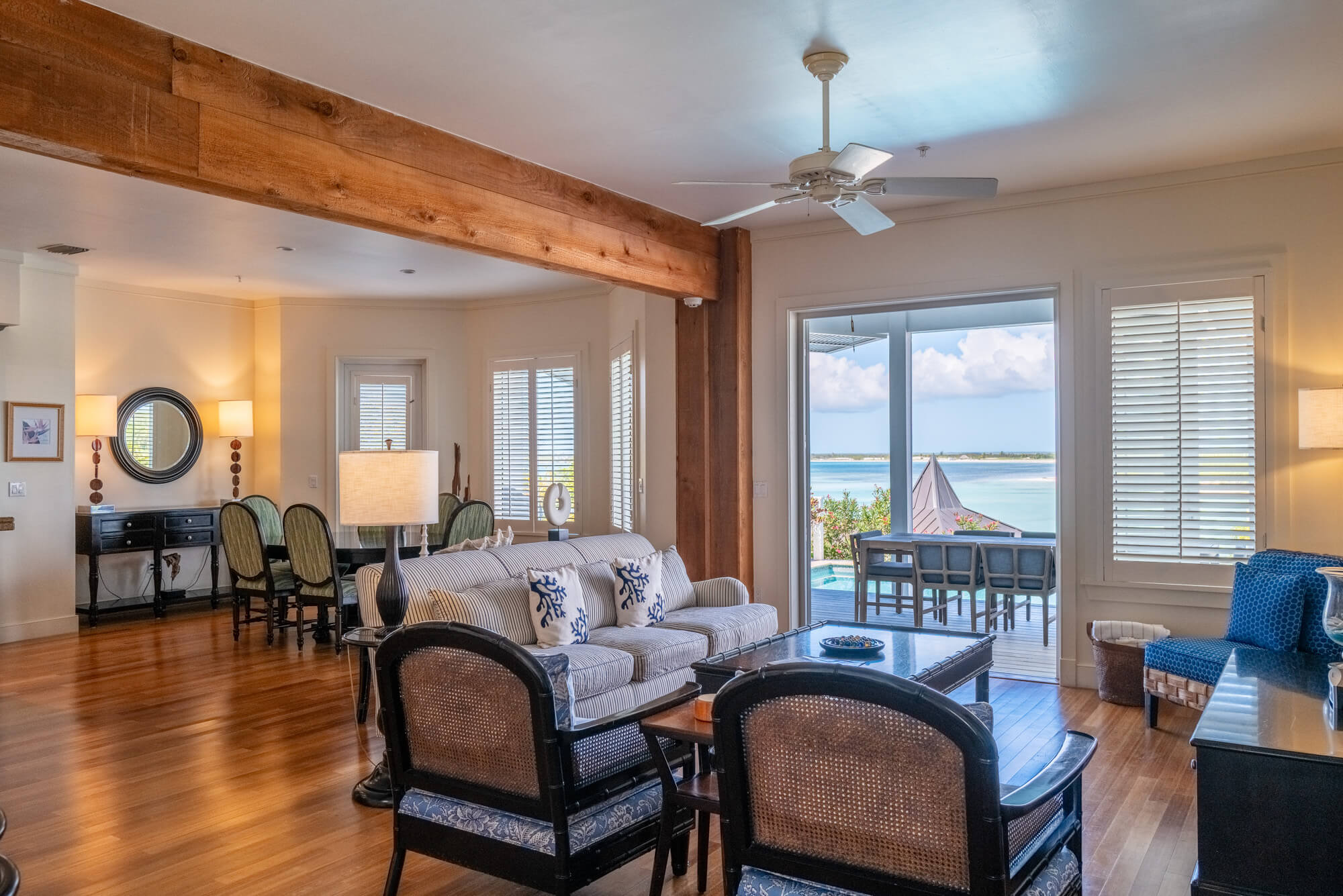 Spacious living room with a large wooden beam, ceiling fan, and island-view windows looking out to the ocean balcony.