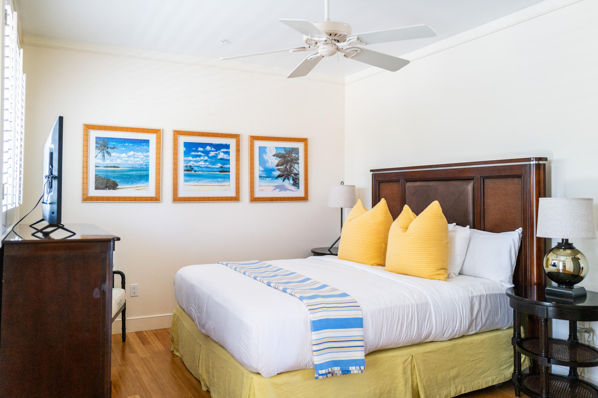 Hotel room with a white bed, yellow pillows, blue striped runner, and a dark wooden headboard under beach-themed framed art on the wall.