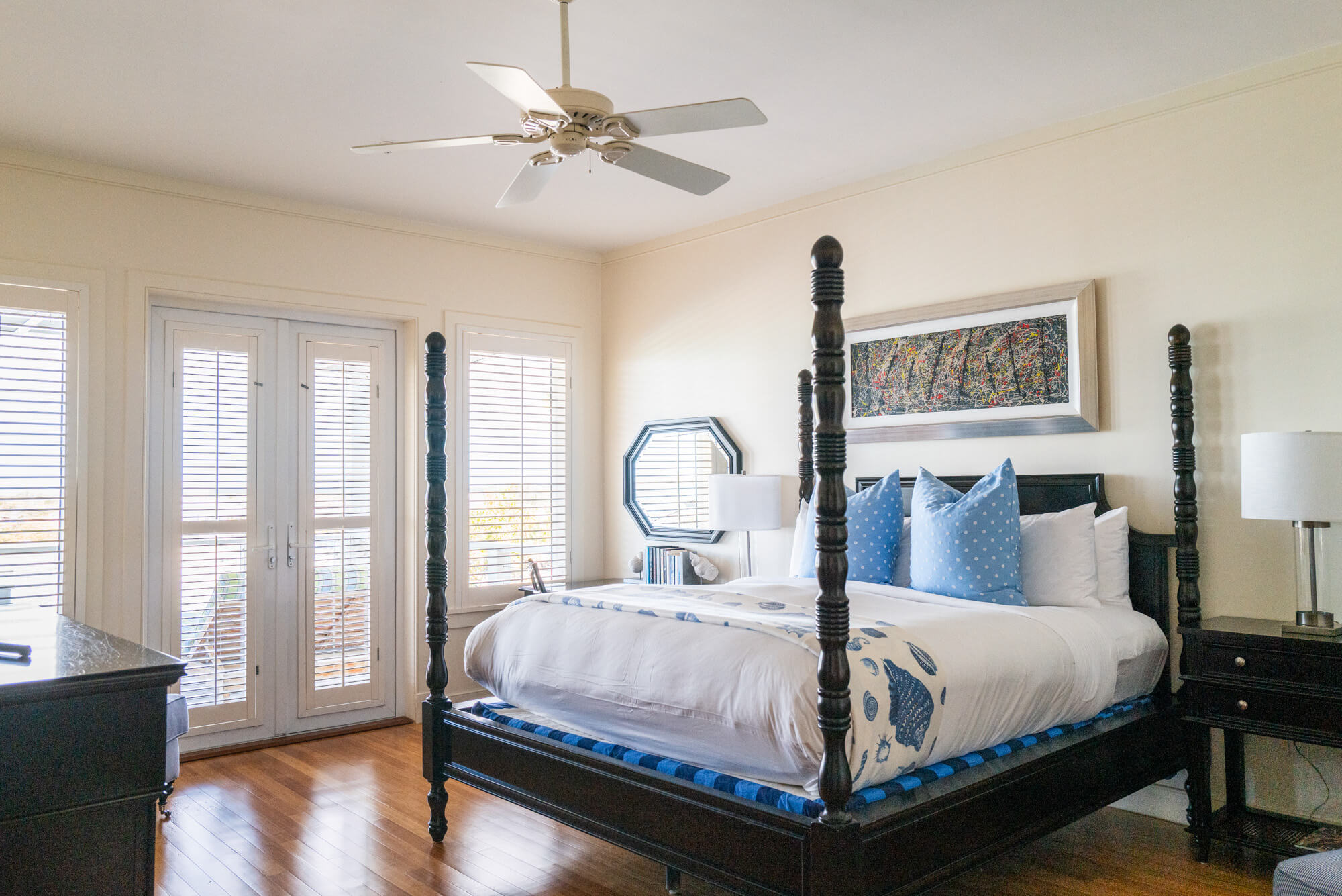 Dark wooden four-poster bed with white and blue bedding in a bright bedroom with blinds on large windows and matching nightstands.