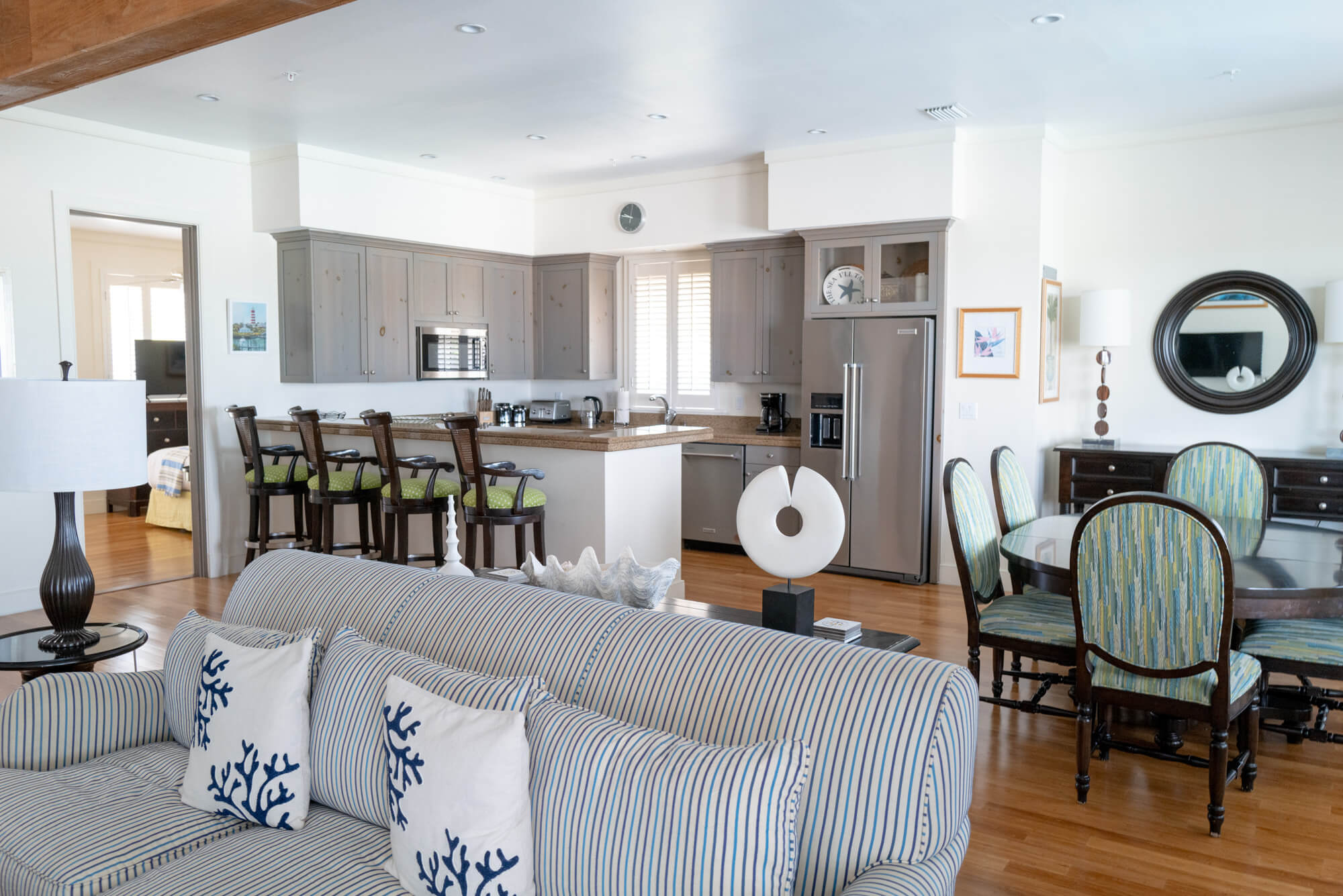 Bright open-plan living area with a blue-striped sofa in the foreground and a modern kitchen with gray cabinets and stainless appliances in the background.