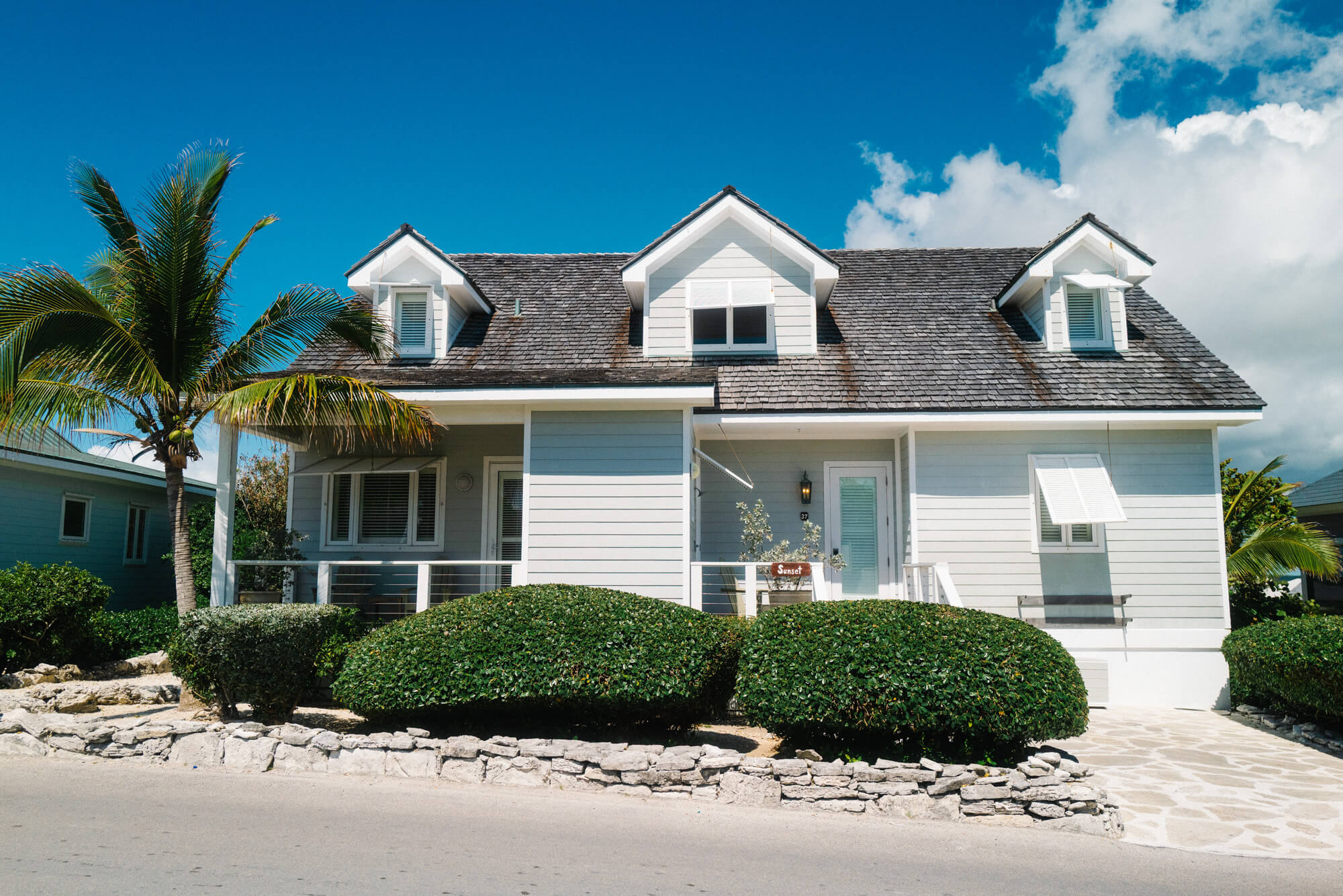 Front view of a light blue coastal house with dormer windows, white trim, and a gray shingle roof under a blue sky with a palm tree nearby