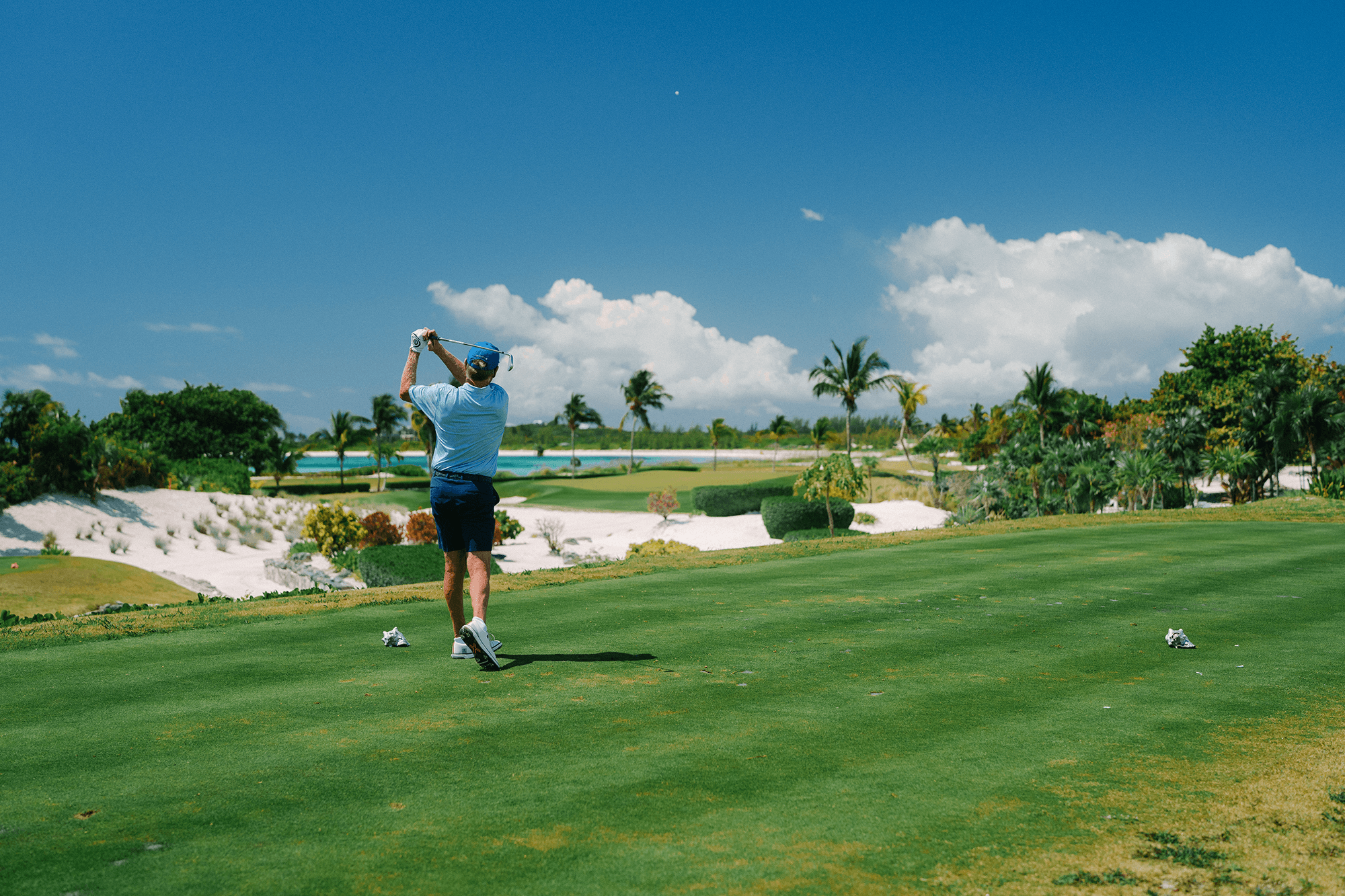 Golfer in blue shirt swinging a club on a sunny golf course with palm trees and sand traps in the background