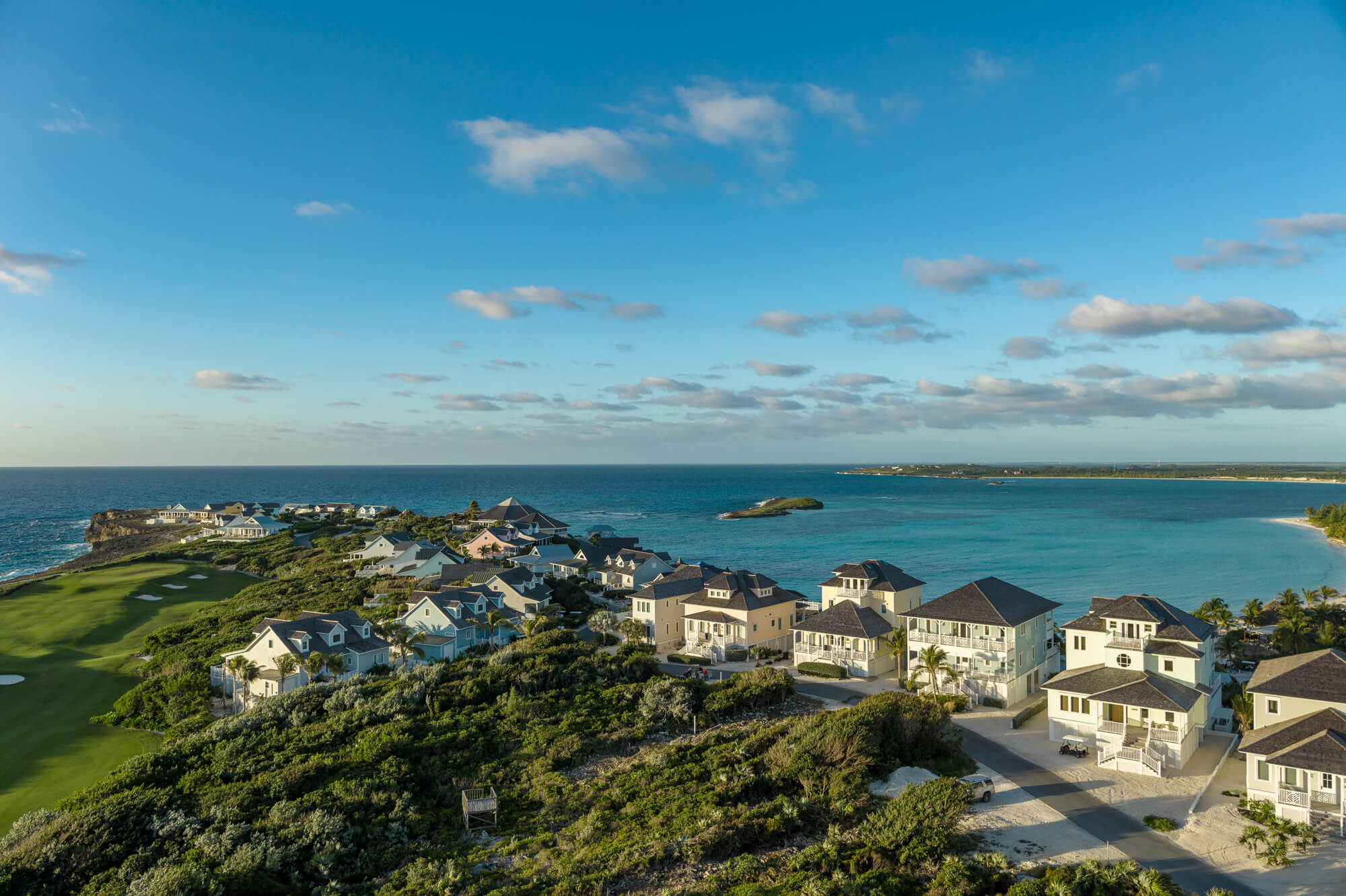 Aerial view of a coastal neighborhood with white houses along the shore, green lawns and shrubbery, and a calm blue ocean under a clear sky.