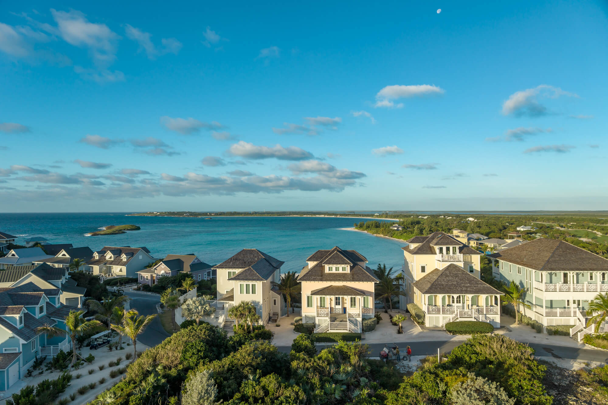 Aerial view of a coastal residential neighborhood with beach houses along a shoreline and the ocean in the distance under a blue sky.