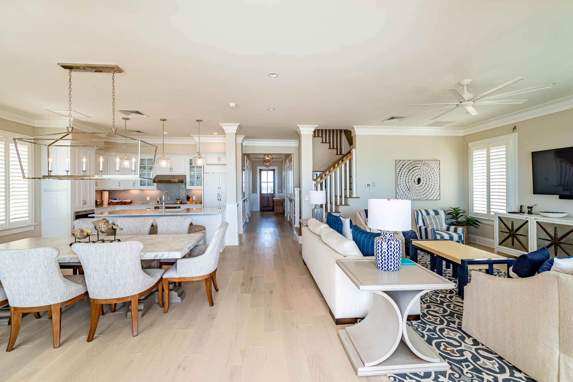 Bright, open-concept living and kitchen area with a long dining table, pendant lights, white cabinets, and a staircase in the background.