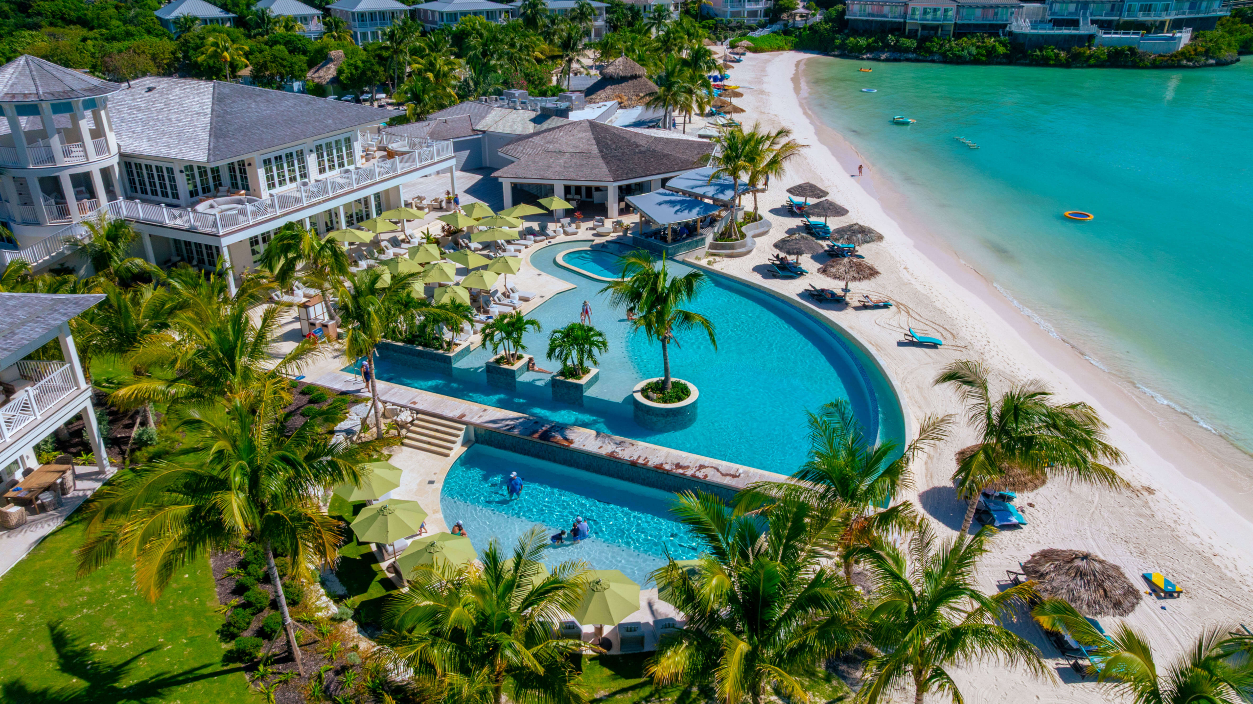 Aerial view of a tropical resort with a curved pool, green umbrellas, palm trees, and a white-sand beach along turquoise water