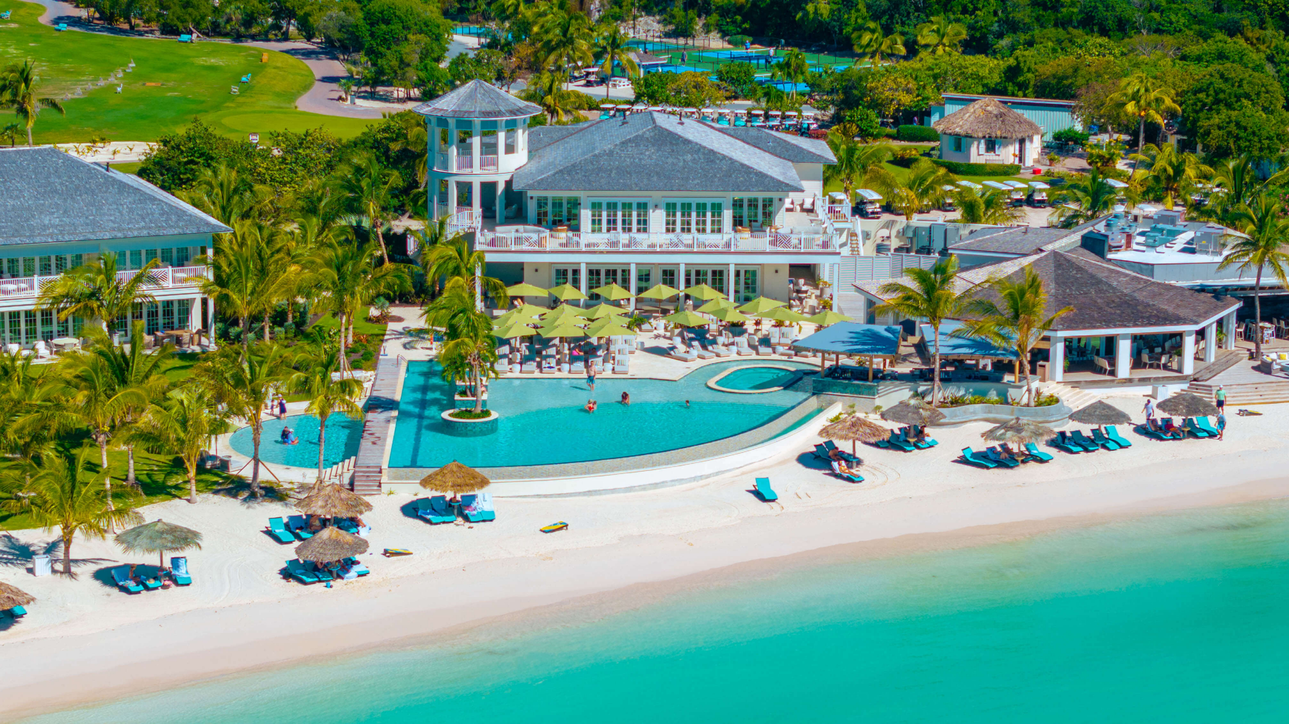 Aerial view of a tropical resort with a large pool area, yellow umbrellas, lounge chairs, palm trees, and white buildings along a sandy beach.