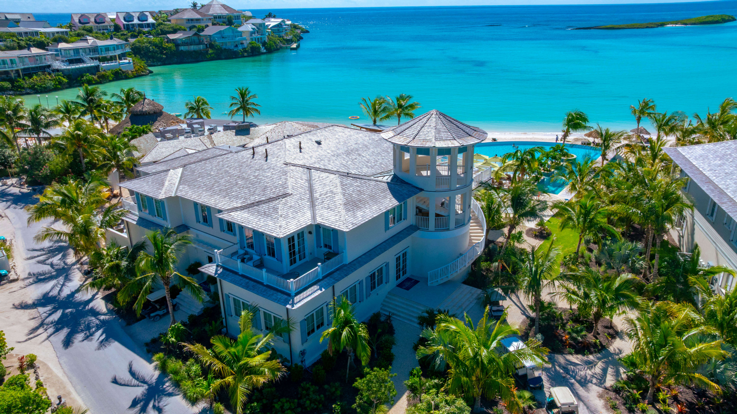 Aerial view of a coastal resort with white villas, palm trees, a circular turret, and turquoise ocean waters.