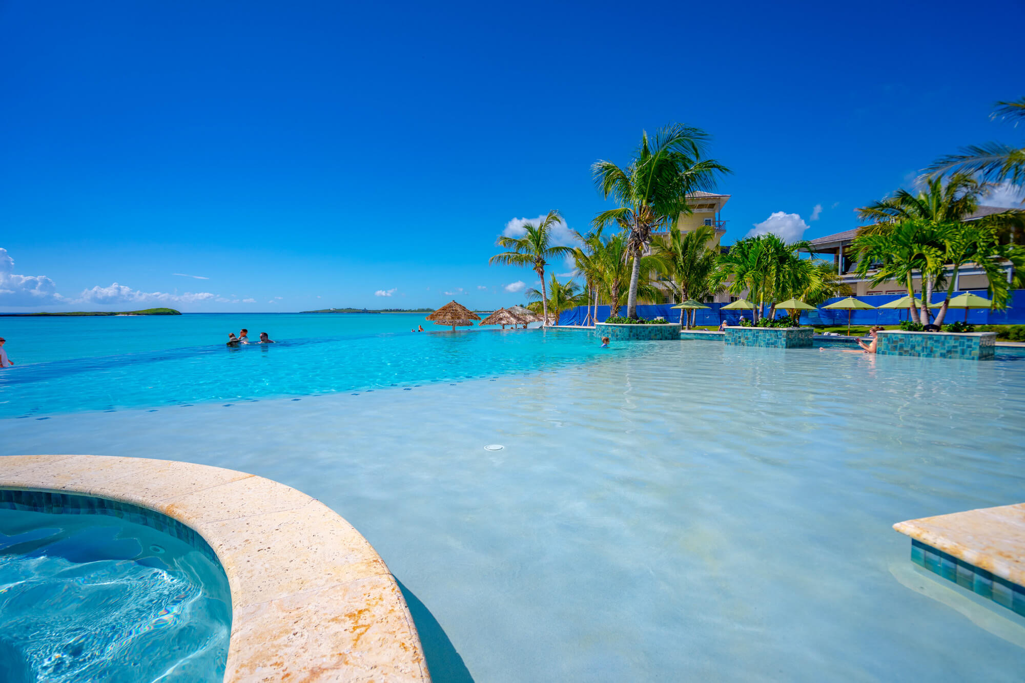 Resort pool with clear blue water, palm trees, thatched umbrellas, and a building in the background under a bright sky