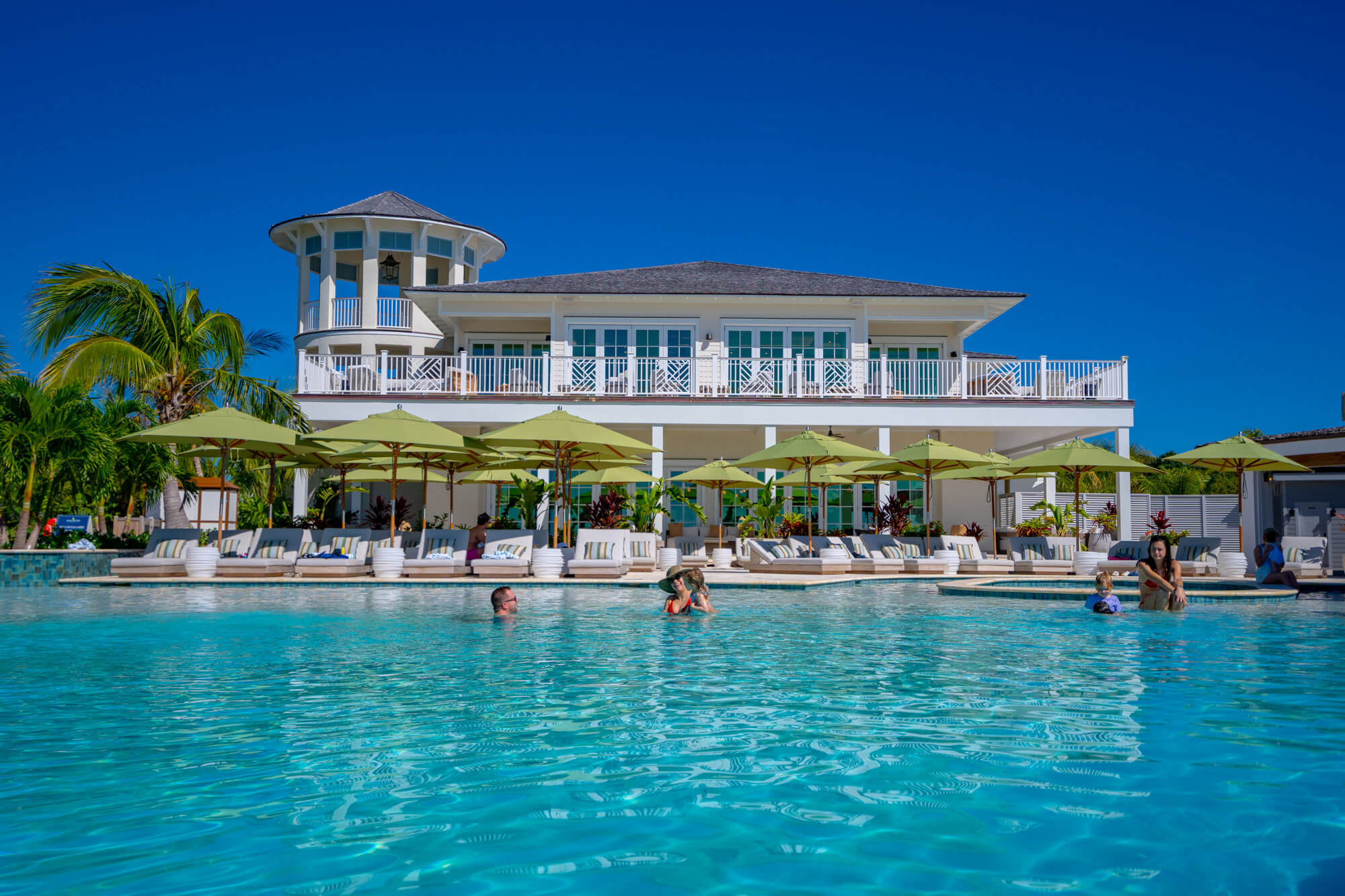 Resort-style pool area with turquoise water, white lounge chairs under green umbrellas, palm trees, and a white multi-level building with balconies in the background under a clear blue sky.