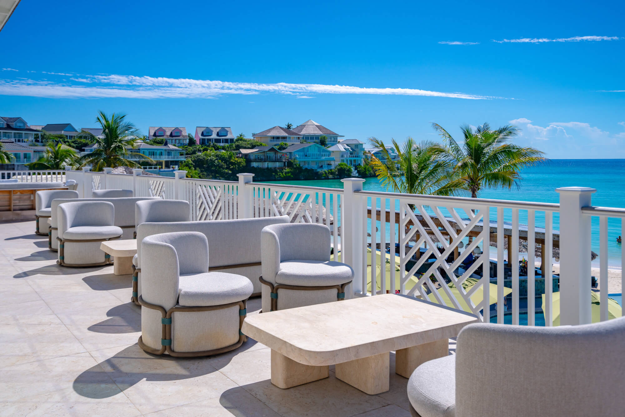 Outdoor resort terrace with white cushioned seating overlooking the turquoise ocean and palm trees.