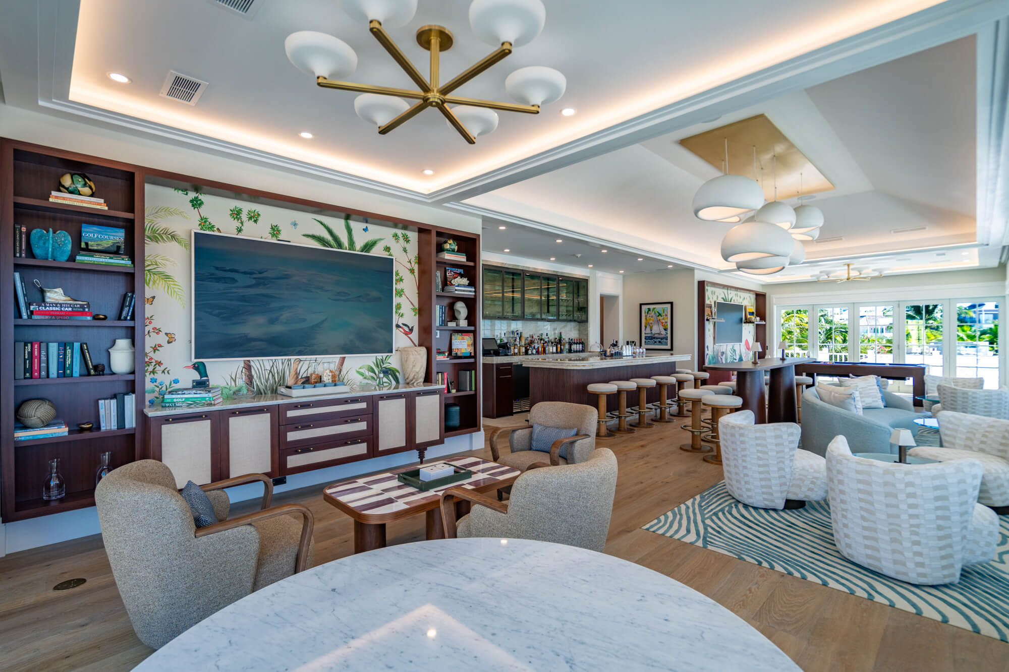 Luxurious tropical-themed hotel lounge with bookshelves, a large TV, and a bar area under warm ceiling lighting.