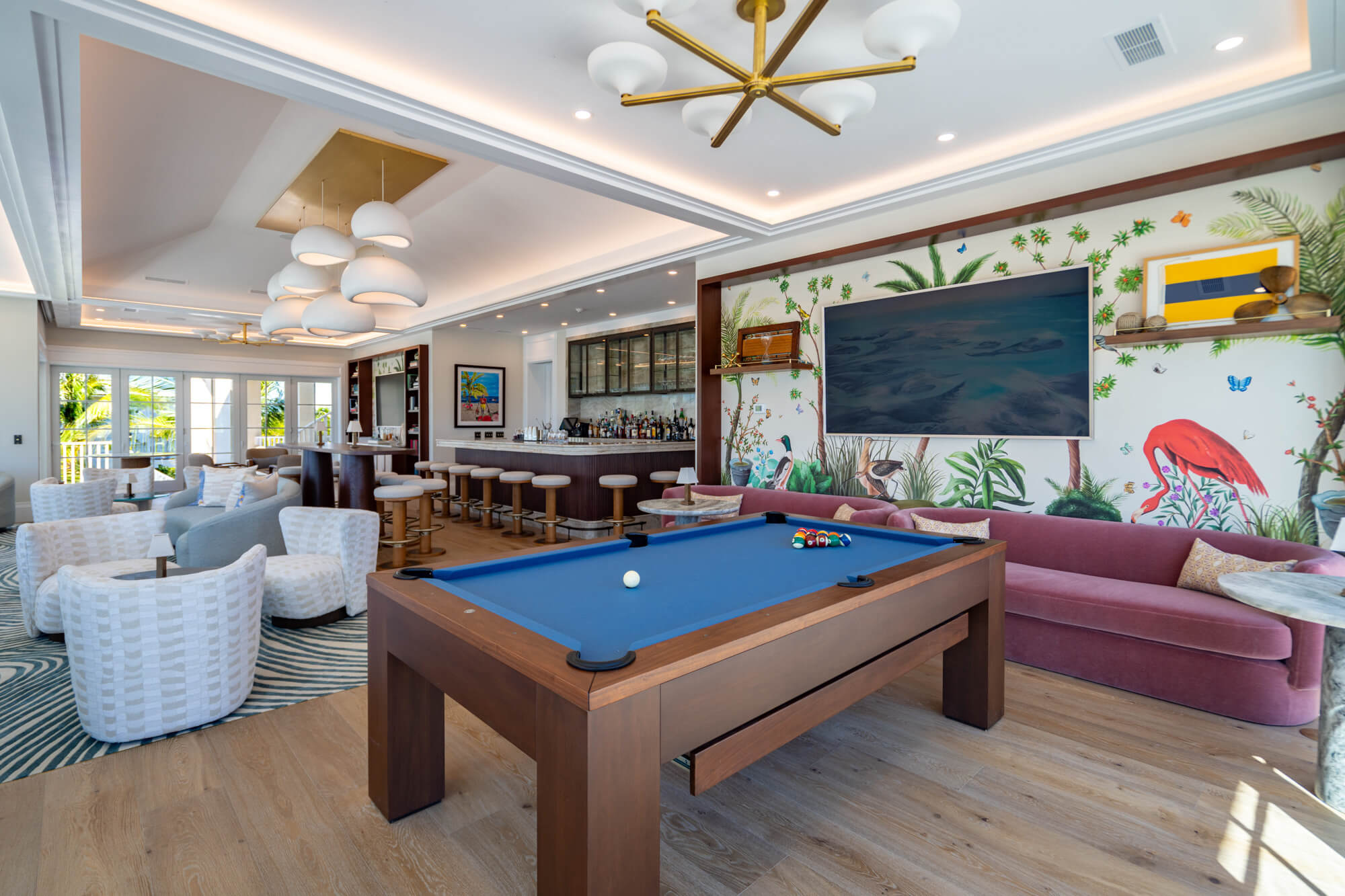 Luxe lounge with a billiard table in the foreground, tropical mural wall, TV, bar seating, and white wicker chairs on wood floors.