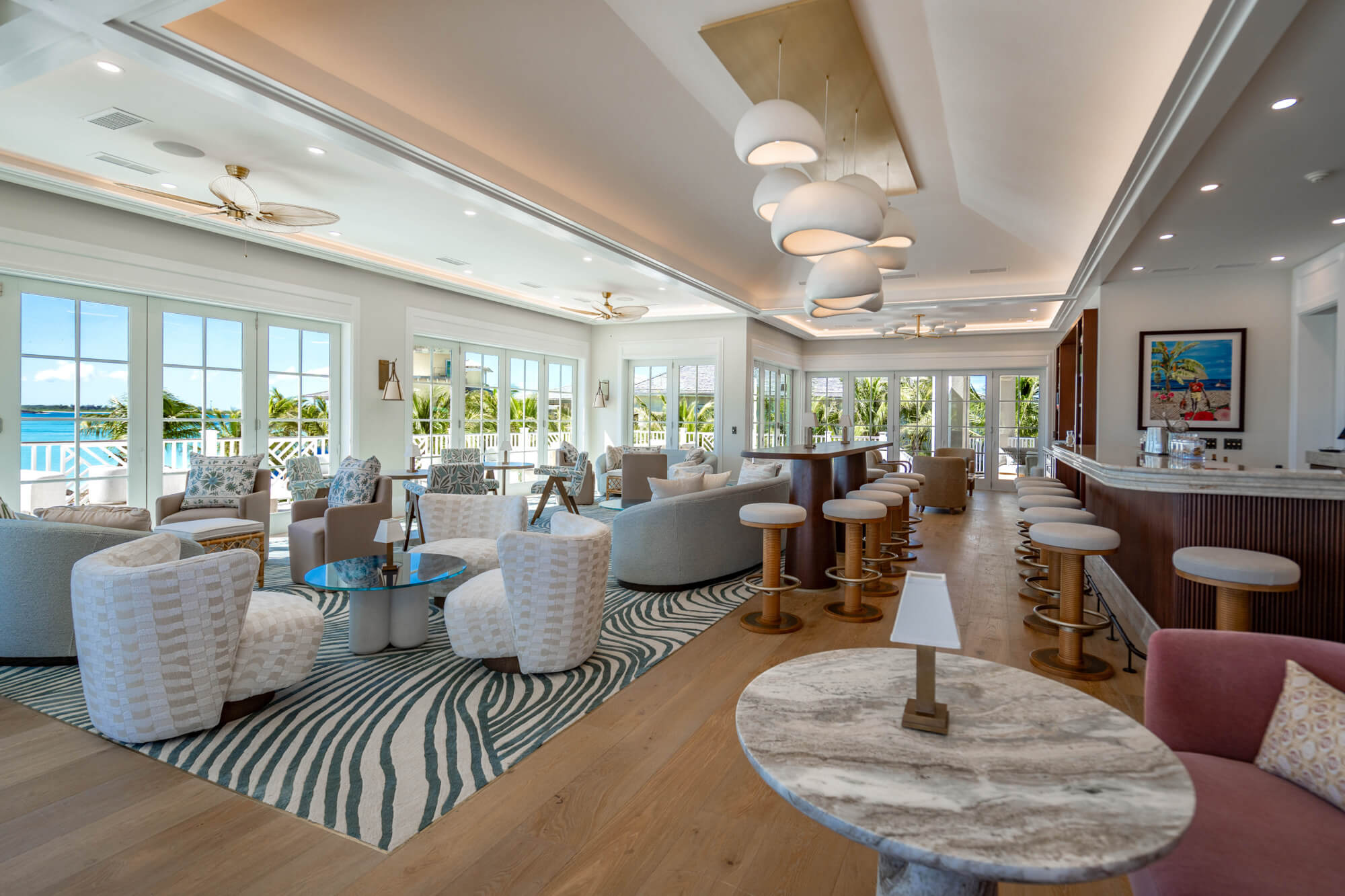 Coastal hotel lounge with plush white and blue seating, round marble tables, and large windows showing palm trees and the ocean outside.