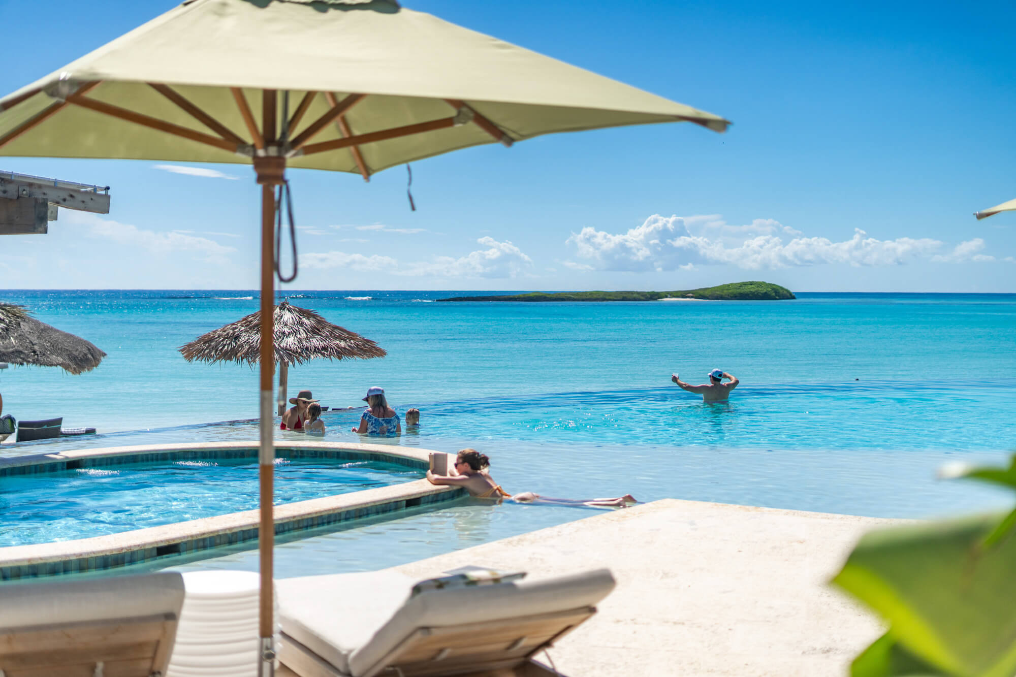 Outdoor resort poolside scene with lounge chairs and large beige umbrellas, people swimming and relaxing under a clear blue sky by the beach.