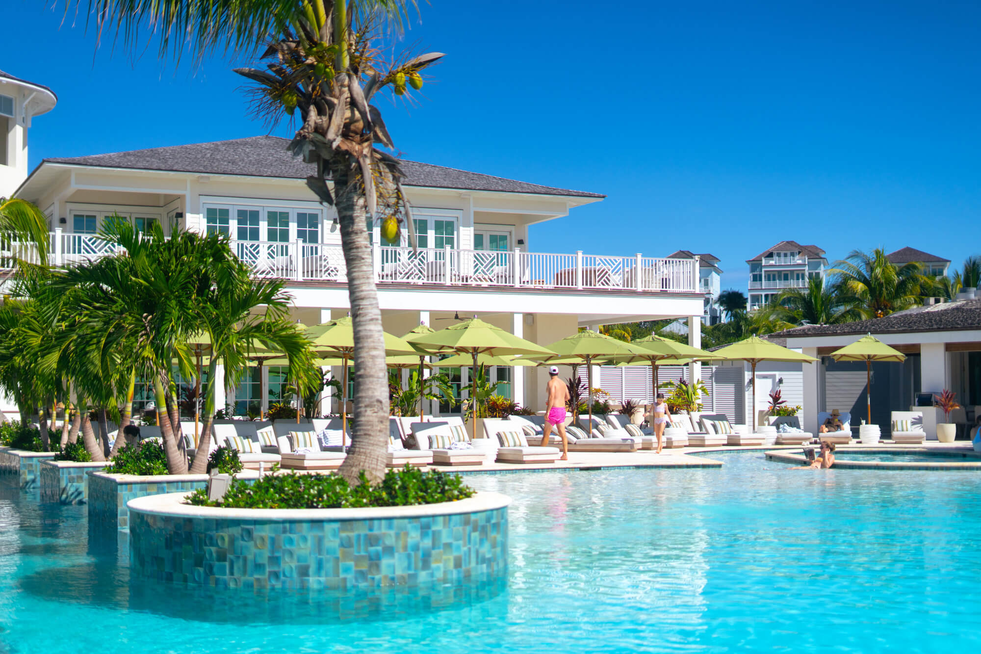 Poolside at a tropical resort with turquoise water, lounge chairs, lime umbrellas, palm trees, and white balconies on the buildings.