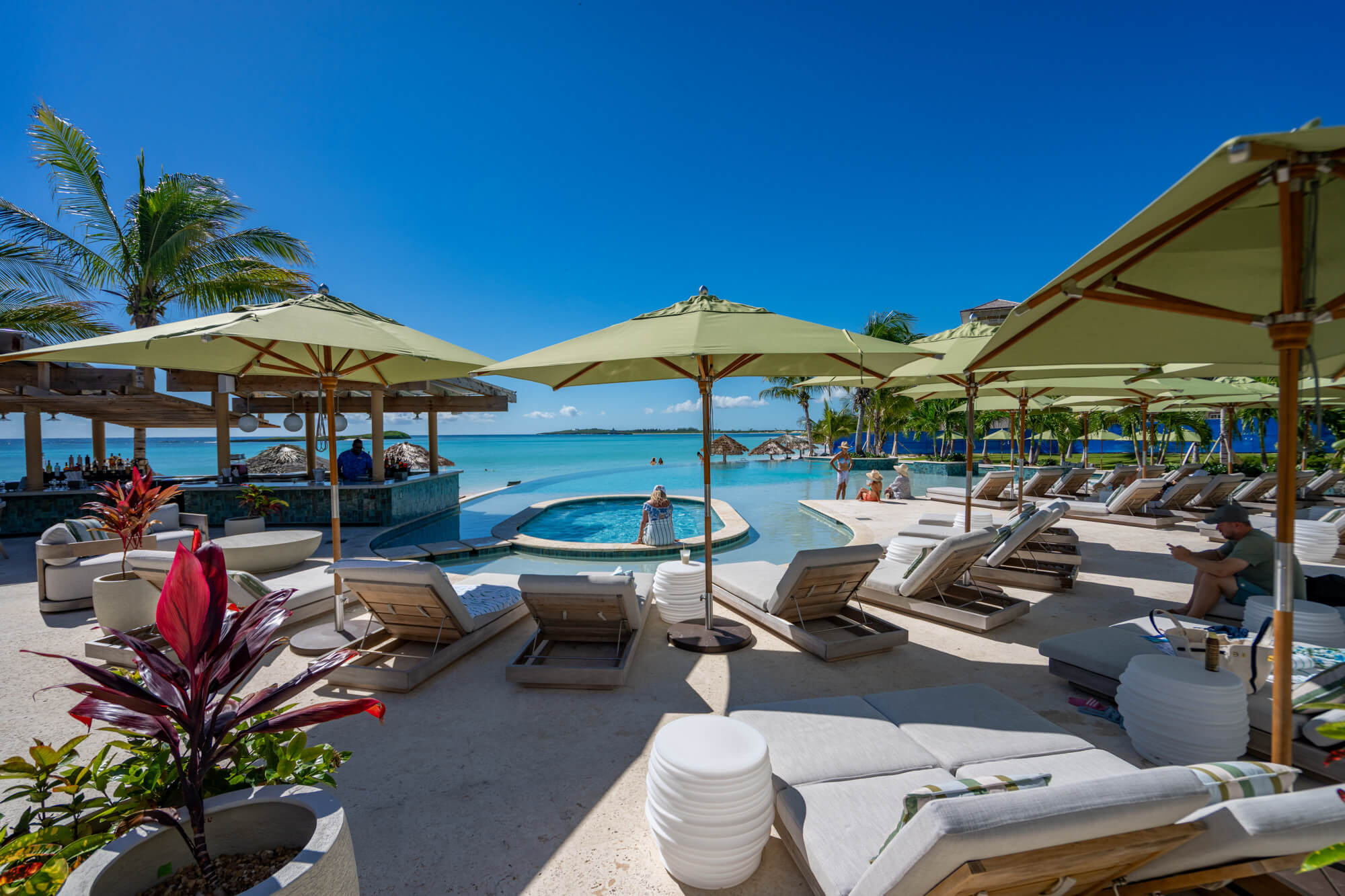 Resort pool area with white loungers, green umbrellas, and palm trees under a clear blue sky