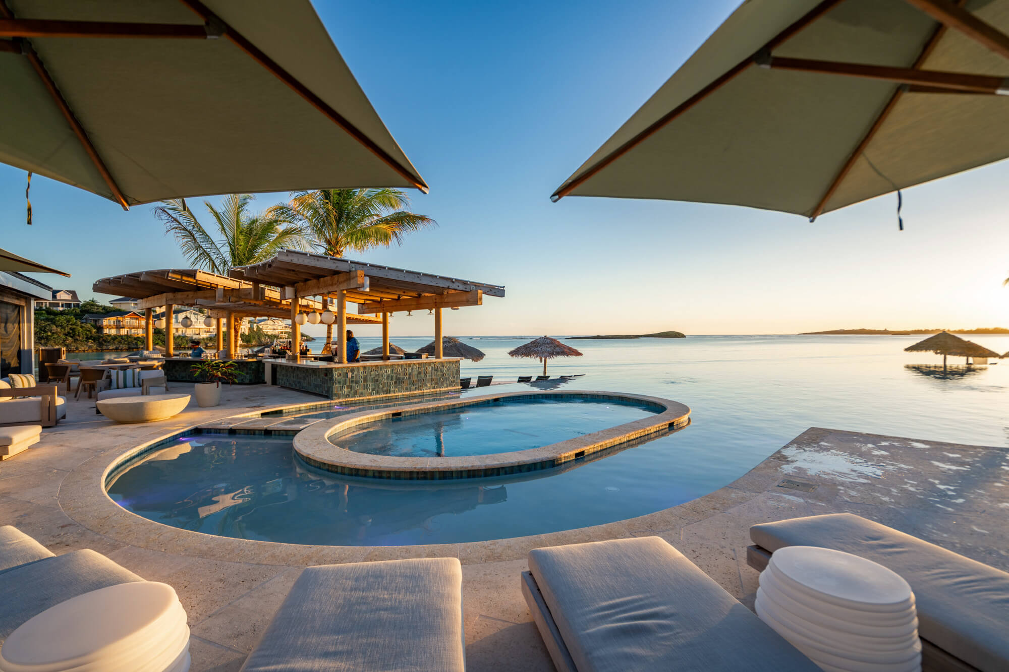 Resort pool area at sunset with lounge chairs, umbrellas, palm trees, and calm sea in the background.