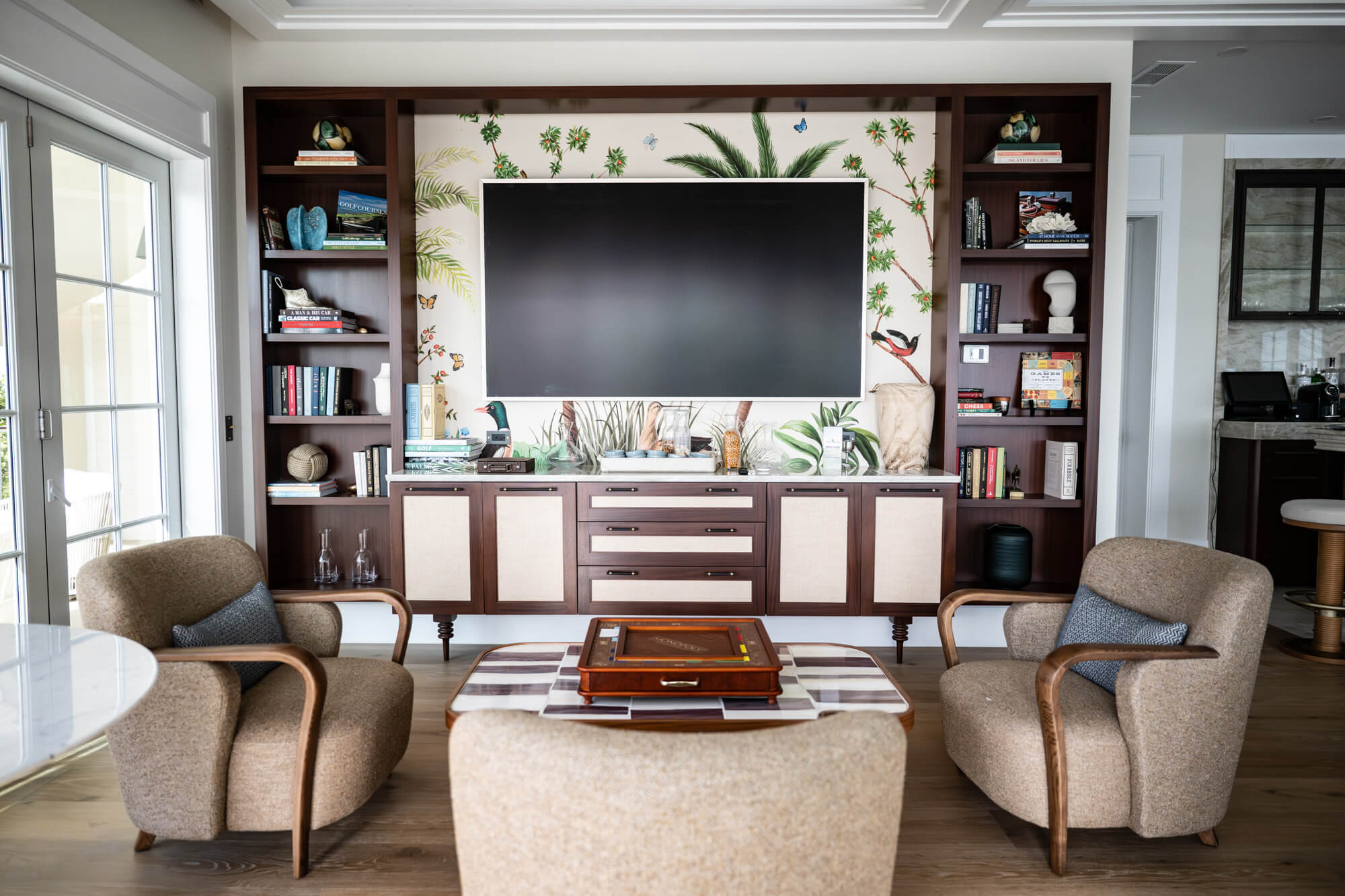 Living room with a large flat-screen TV centered in a tropical wallpaper wall, flanked by dark wood shelves filled with books and decor.