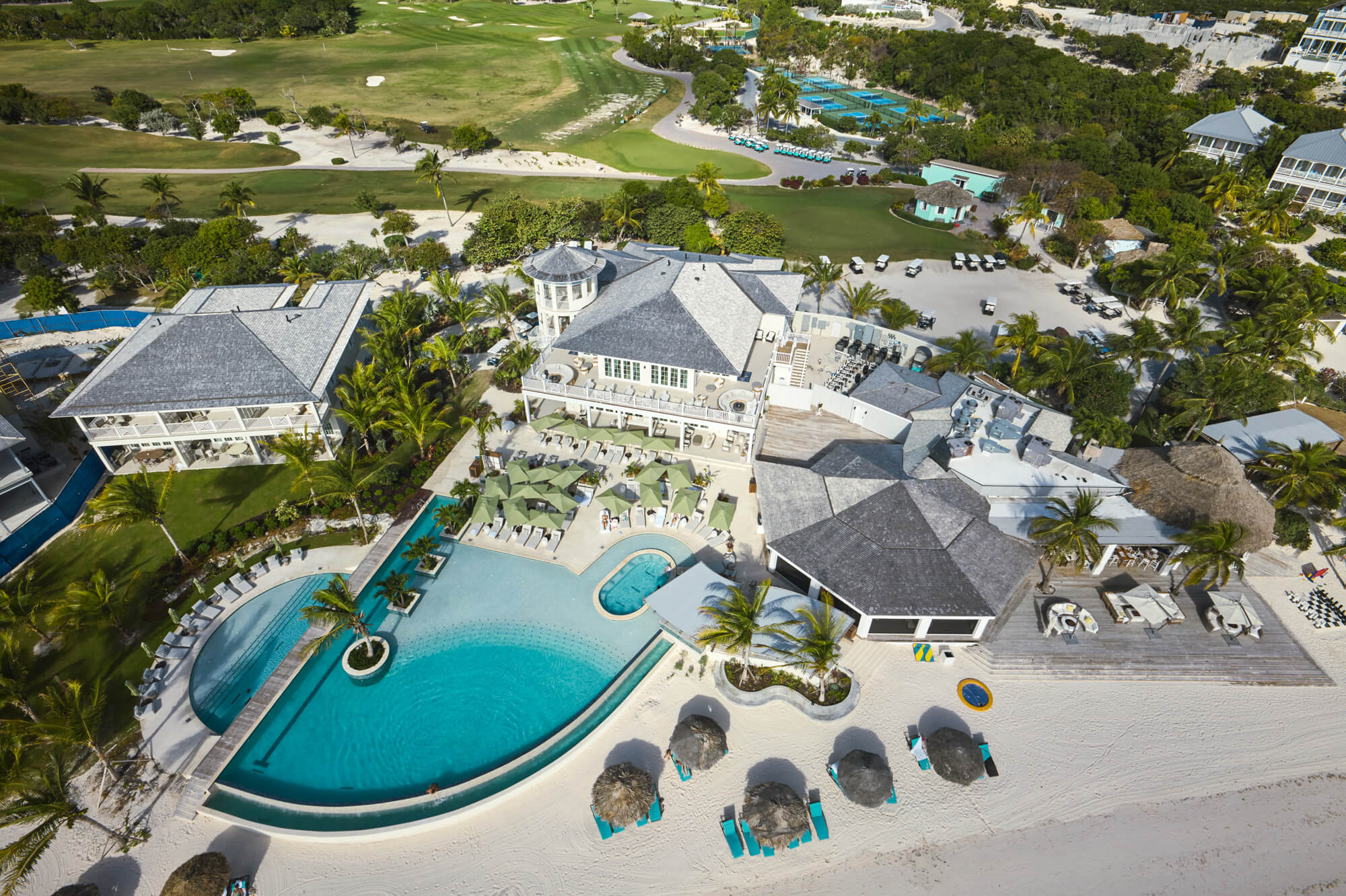 Aerial view of a tropical resort with a curved outdoor pool, white buildings, palm trees, and a sandy beach.