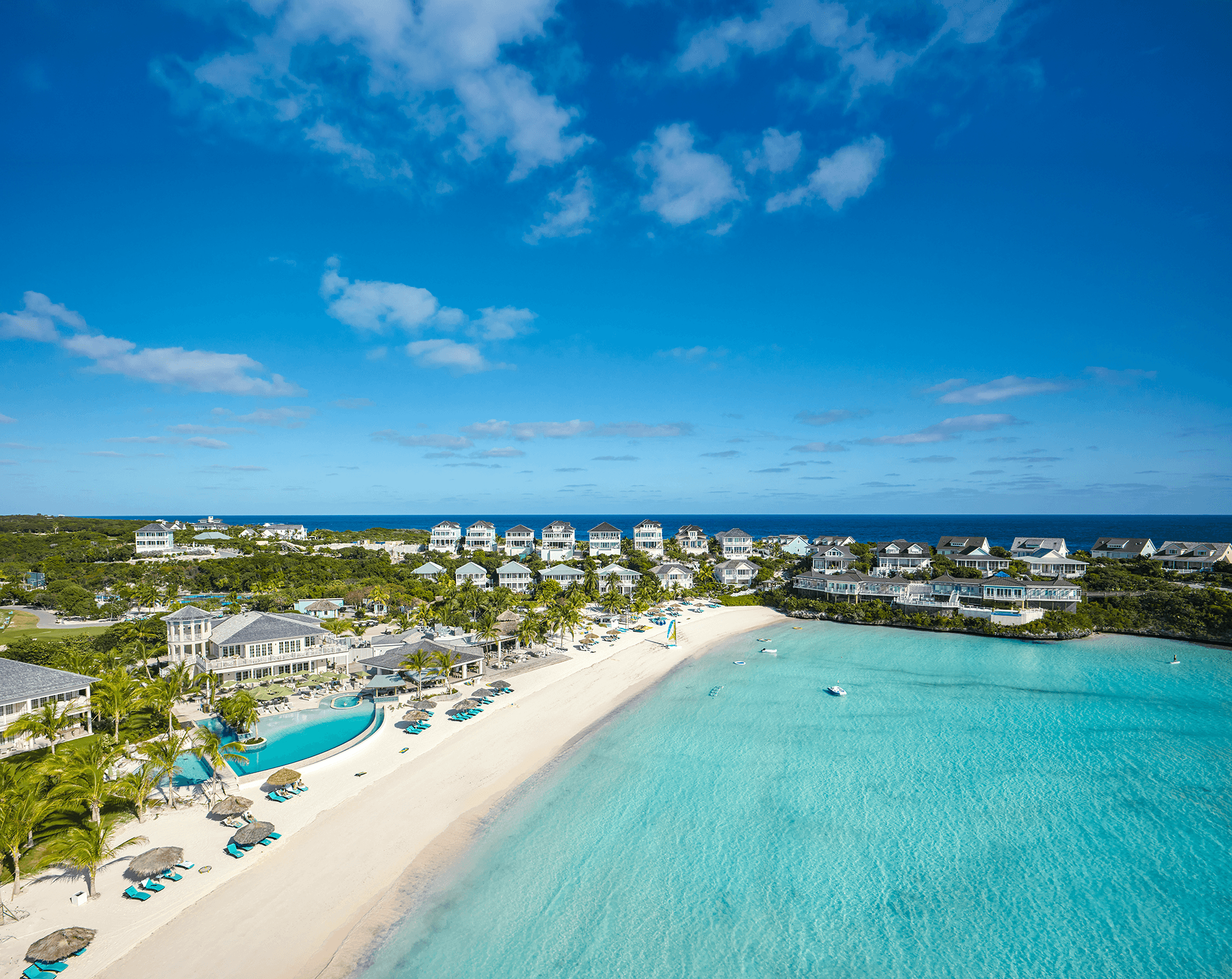 Seaside resort with white-sand beach, turquoise water, and row of beach-front villas under a bright blue sky.