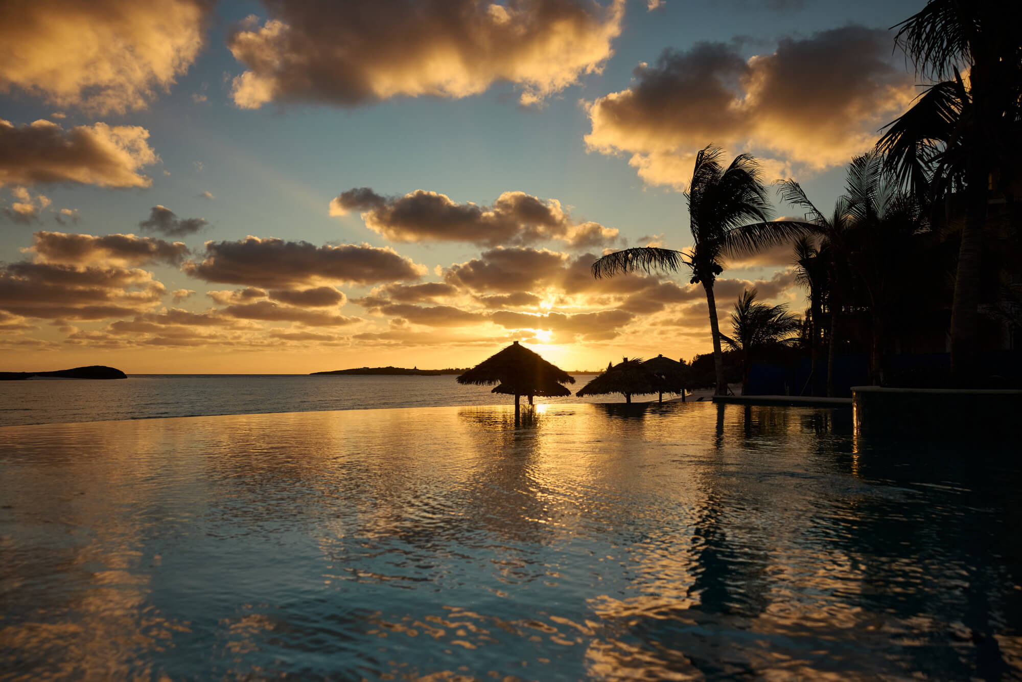 Golden sunset over calm waters with silhouetted palm trees and thatched umbrellas along the beach, reflecting in the foreground pool.