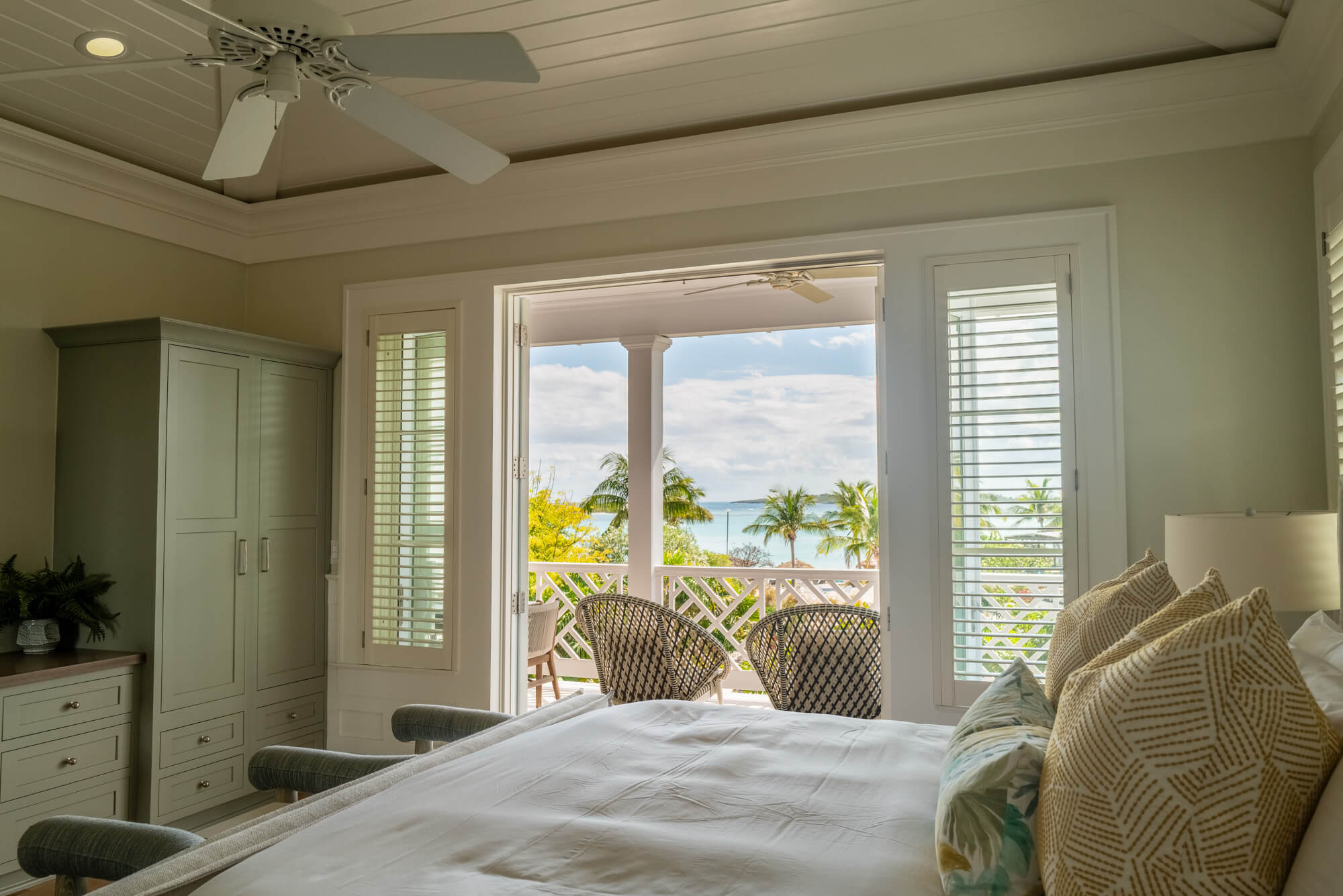 Bright bedroom with open French doors to a sunny balcony, tropical ocean view beyond palm trees, and a white ceiling fan above.