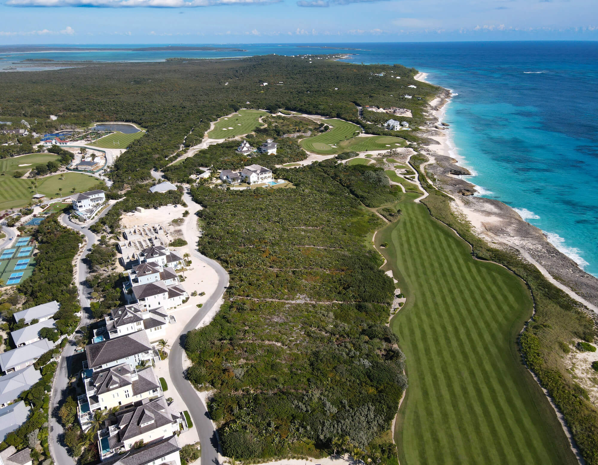 Aerial view of a coastal resort with a winding golf course, white houses, and turquoise sea along a rocky shoreline.