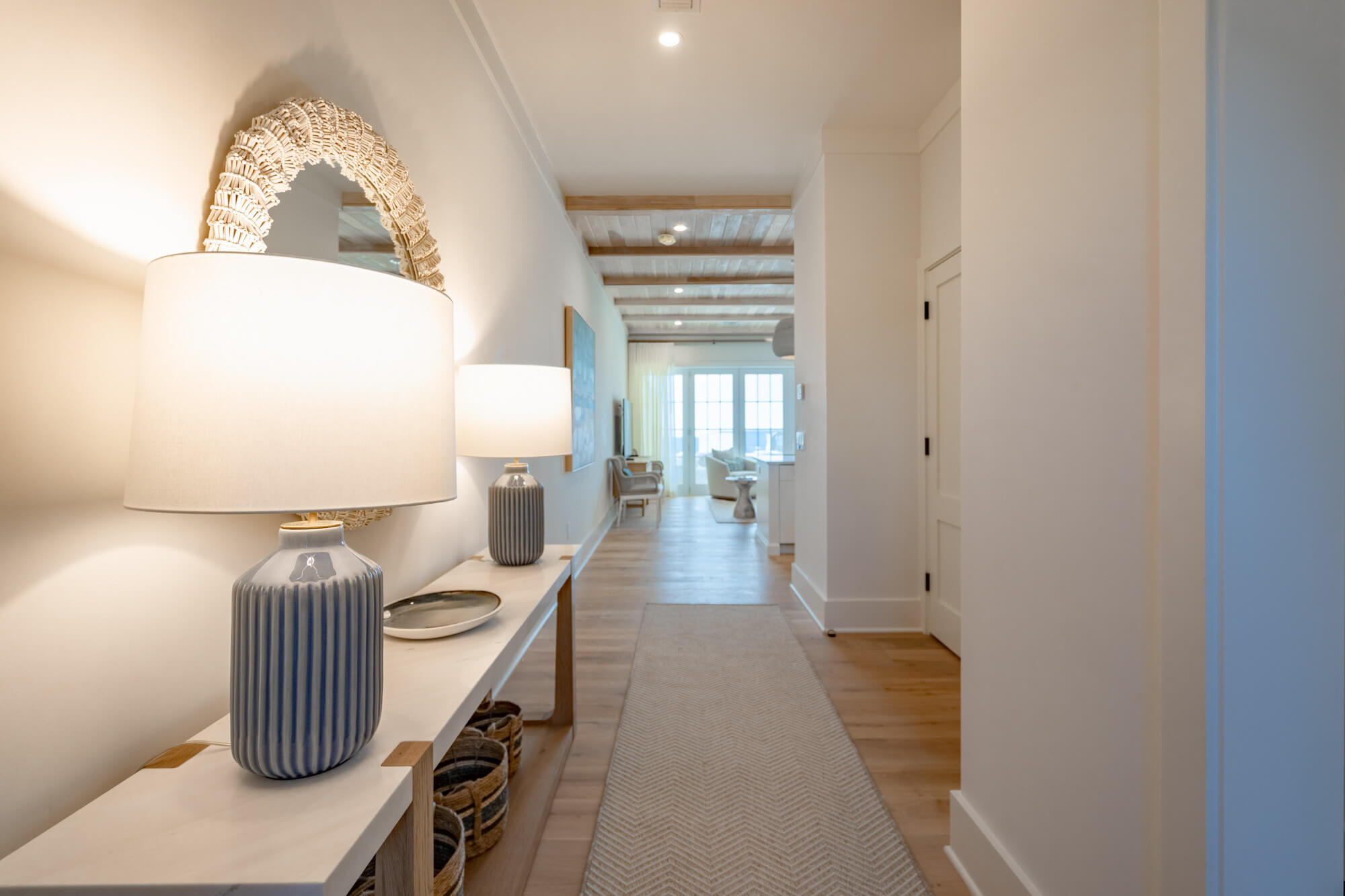 Hallway with a white console table, striped ceramic lamps, and wicker baskets, leading to a sunlit living area.