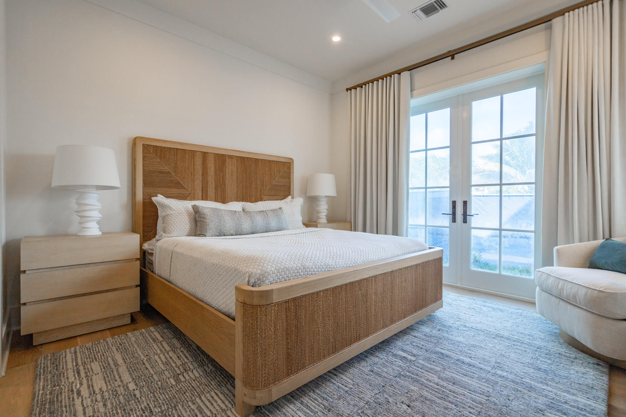Bright bedroom with a light wood bed, matching nightstands, white lamps, and large glass doors framed by cream curtains. Outside view shows greenery.