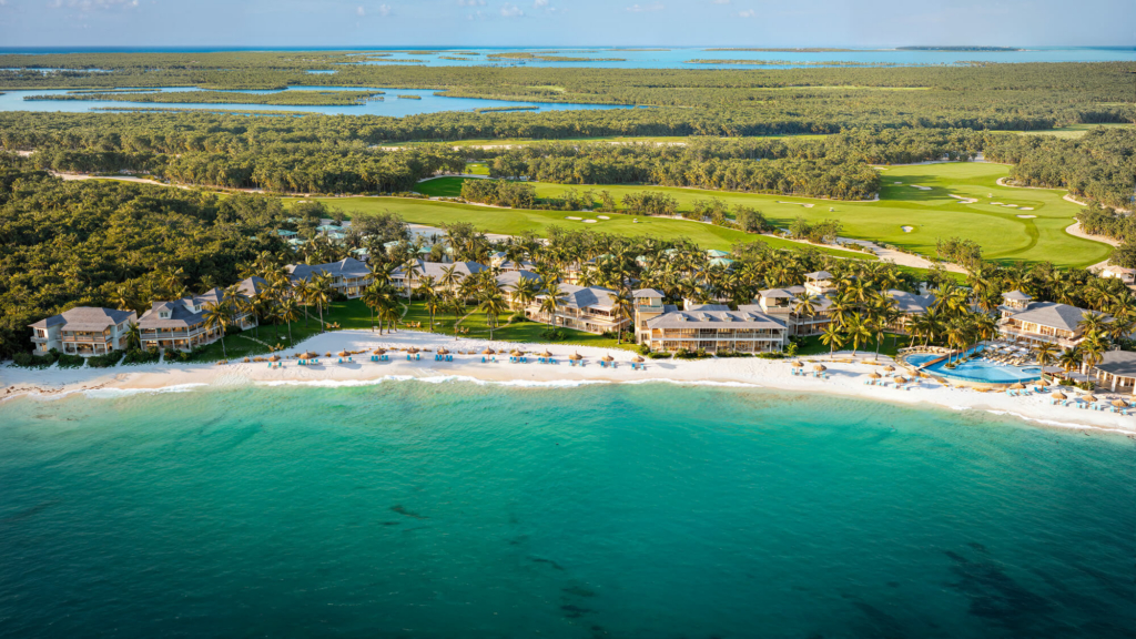 Aerial view of a tropical resort with beachfront villas, palm trees, and a golf course beside turquoise water.