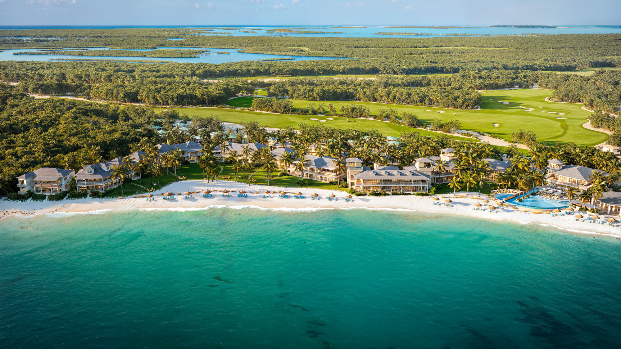 Aerial view of a tropical resort with beachfront villas, palm trees, and a golf course beside turquoise water.