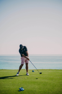 Golfer mid-swing on a seaside golf course, wearing a cap, dark shirt, and pink shorts, with the ocean in the background and a golf club extended toward the ball.",