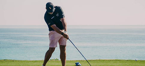 Golfer in a dark shirt and pink shorts swinging a club on a coastal golf course with the sea in the background